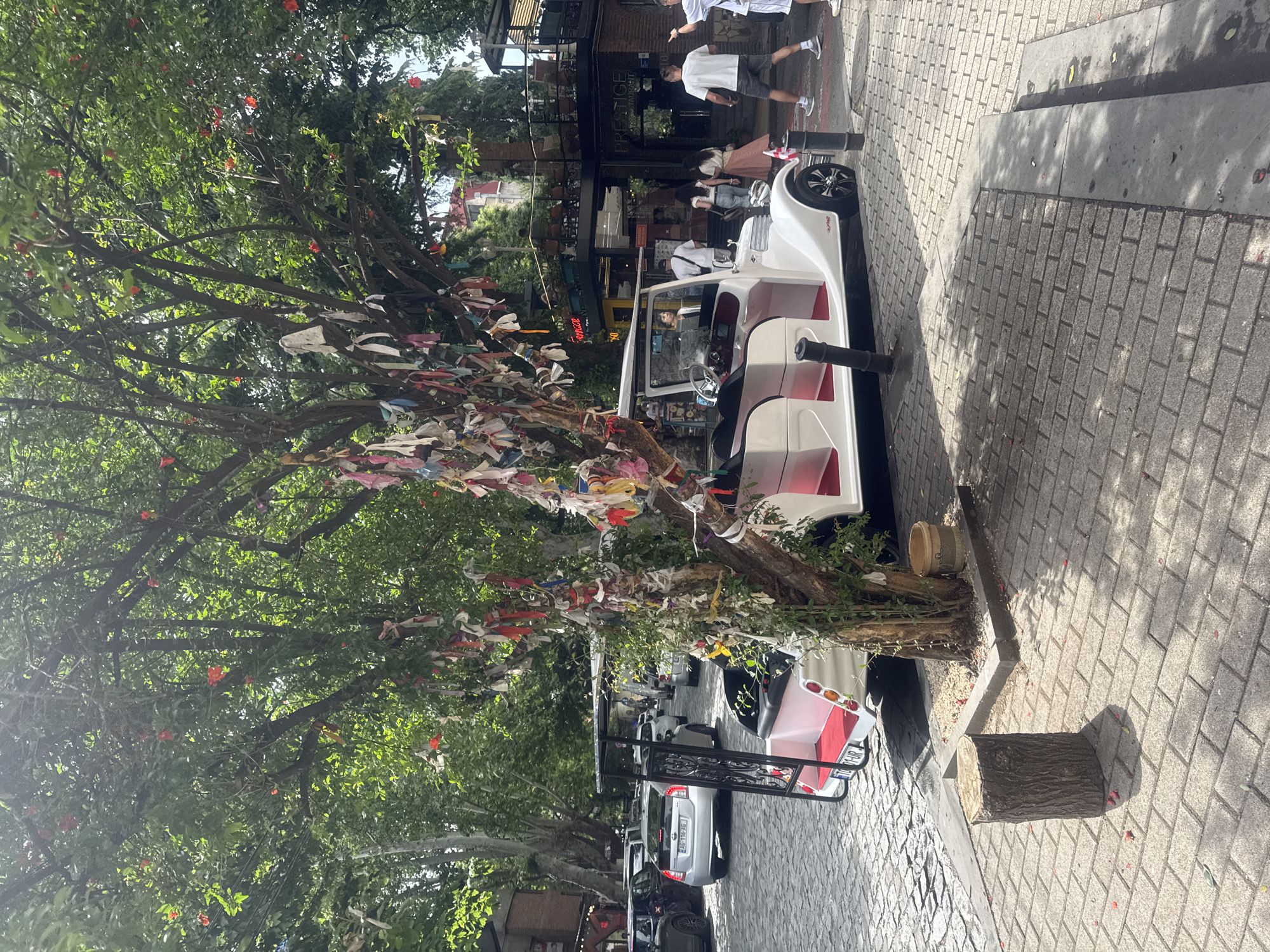 An outdoor scene in Tbilisi, Georgia, features a white and red open-air vehicle, similar to a golf cart, parked on a cobblestone street. The vehicle has multiple rows of seating and a black canopy. Adjacent to the vehicle, a tree is adorned with numerous colorful ribbons tied to its branches, indicating a cultural or religious tradition. In the background, several other vehicles are parked along the street, and buildings with shops and restaurants are visible. People are walking along the sidewalk, some appearing to be tourists. The overall atmosphere suggests a lively urban environment, likely a tourist area within the city.