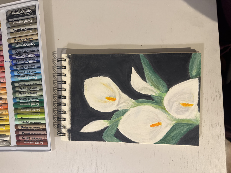 The scene, located in Mexico City, Mexico, depicts an art setup on a light-colored, textured surface, likely a desk or table. A vertically oriented, spiral-bound sketchbook is open, showcasing an oil pastel drawing of four white Calla Lilies. Each lily possesses an orange spadix and is accompanied by green foliage, all rendered against a dark, possibly black, background. One lily is depicted as an unopened bud. Positioned above the sketchbook is a white plastic case containing a complete set of "Pentel Oil Pastels," with numerous pastel sticks arranged in color-graded rows. Shadows from the pastel case and the sketchbook's spiral binding are visible on the underlying surface. No individuals or active interactions are present.