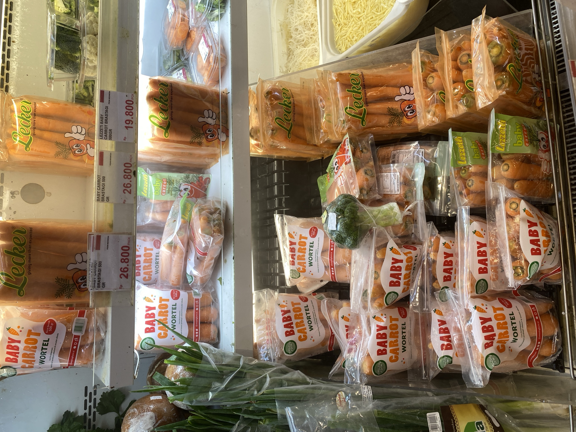 The image shows a refrigerated display case at a grocery store in East Jakarta, Indonesia, filled with various packaged baby carrots. The products are predominantly from brands labeled "Lecker" and "Amazing Farm," with some appearing to be from "Lezzetti Baby." The carrots are packaged in clear plastic bags and some in clear plastic containers. The display case is well-lit, and the products are neatly arranged. Some of the packaging displays nutritional information and branding in Indonesian. In the foreground, there are also packages of green onions. In the background, other vegetables like broccoli and cauliflower can be seen in clear plastic containers. The overall environment suggests a typical supermarket produce section.