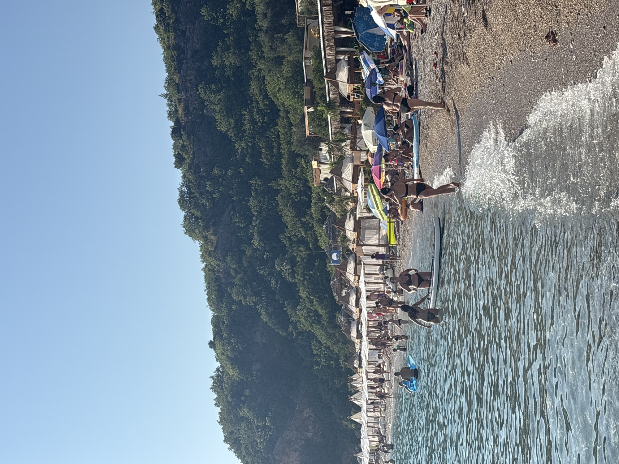 The image depicts a crowded beach scene in Drobnići, Montenegro, under a clear blue sky. The foreground shows the clear, shallow waters of the Adriatic Sea lapping against a pebble beach. Several people are in the water, some wading, others swimming or on inflatable toys. A woman in a bikini is walking out of the water, and another woman is standing on a paddleboard. The beach itself is lined with numerous white cabanas with thatched roofs, offering shaded seating areas. Beach umbrellas in various colors, including blue, green, and pink, are scattered across the sand. Many people are seated under the umbrellas or in the cabanas, sunbathing or relaxing. In the background, a steep, densely forested hillside rises, providing a verdant backdrop to the beach resort. Buildings, possibly part of the resort, are visible nestled amongst the trees and cabanas. The overall impression is of a busy, sunny day at a popular beach destination.