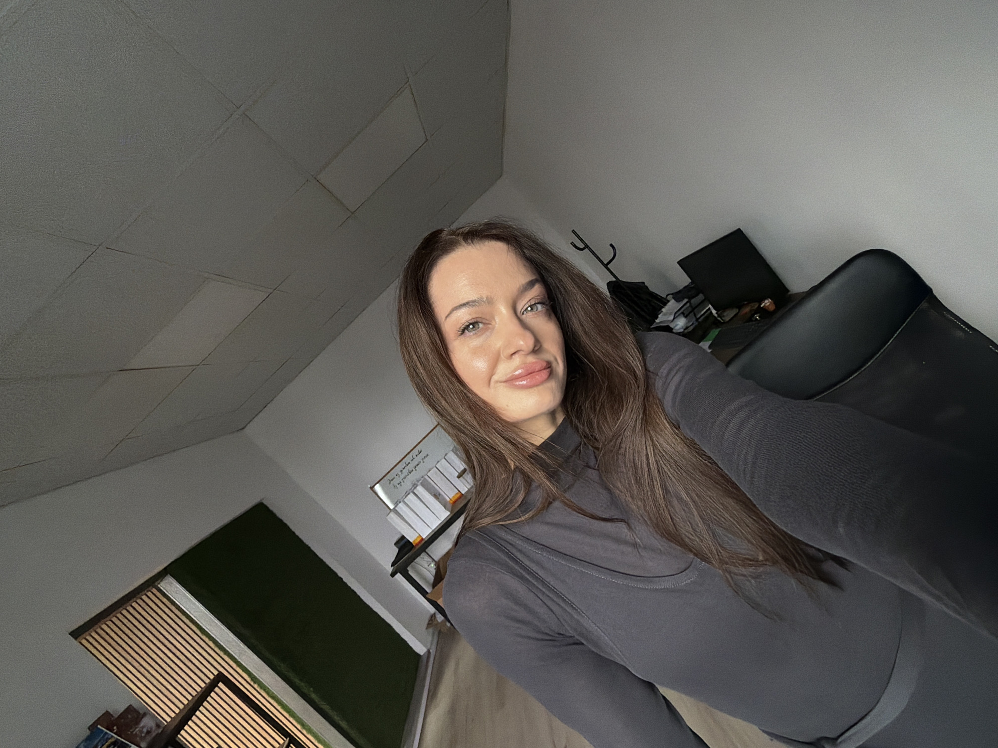 A woman is taking a selfie in an indoor space. The space appears to be an office, with a drop ceiling and a partially visible window with blinds and green screen visible in the lower left. There is a shelf with various objects in the background, and a black chair to the right. The woman has long brown hair, fair skin, and is wearing a grey long-sleeved top. Her expression is slightly smiling. The location is likely inside a building, but it is not possible to specifically identify the location as being in Varna, Bulgaria, from this image alone.
