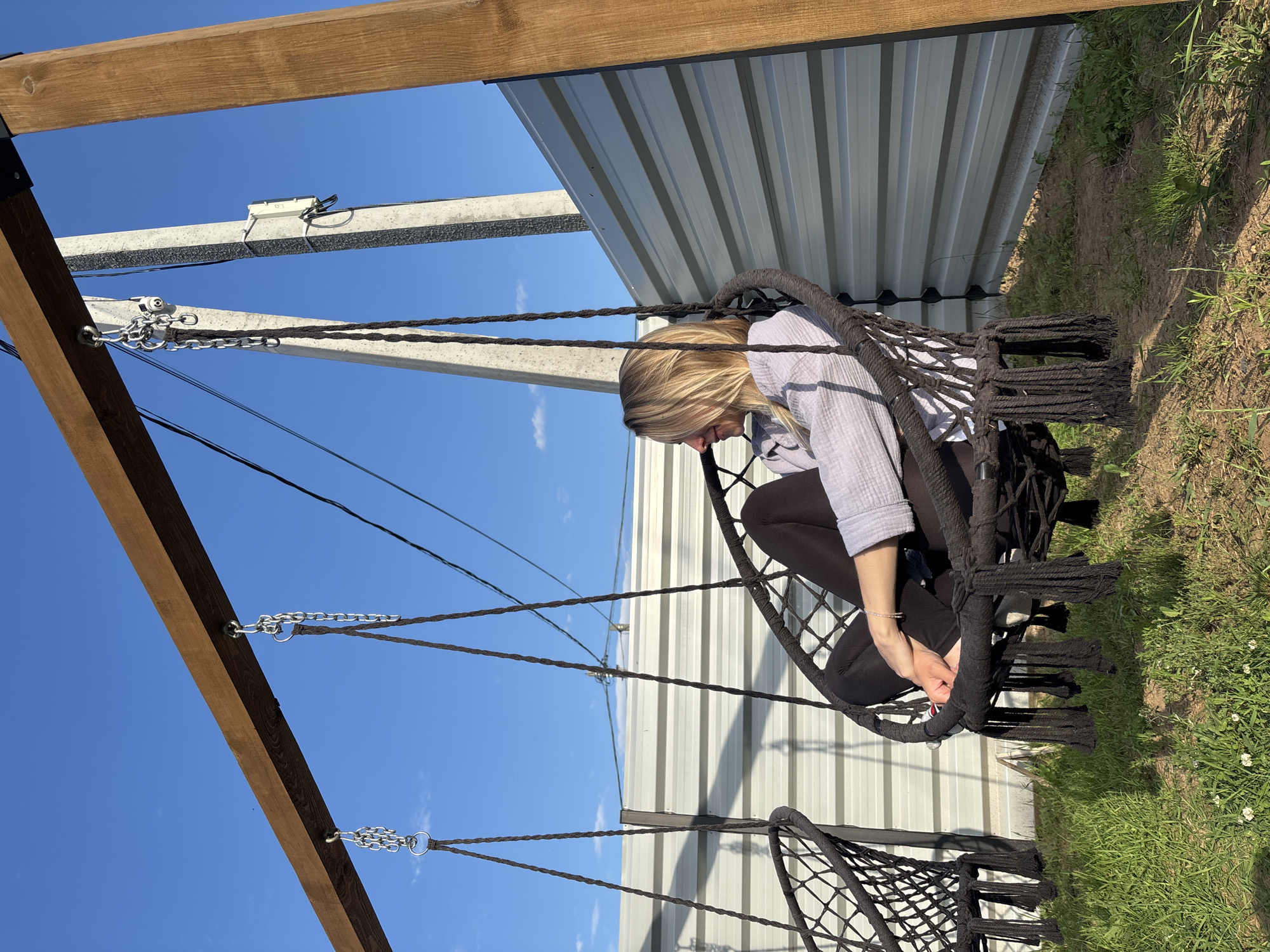 A blonde woman with fair skin is sitting in a black woven swing chair outdoors. She is wearing a light-colored, long-sleeved shirt and black pants. The swing chair is suspended by chains from a wooden beam. To the left, another identical swing chair is visible, also empty. In the background, a corrugated metal fence runs horizontally, with a utility pole with electrical wires situated behind it. The ground is covered in grass and dry soil. The sky is clear and blue with a few wispy clouds. The scene appears to be set in a yard or outdoor recreational area, possibly in the city of Chaikovsky, Russia, given the presence of utility poles and the overall aesthetic.