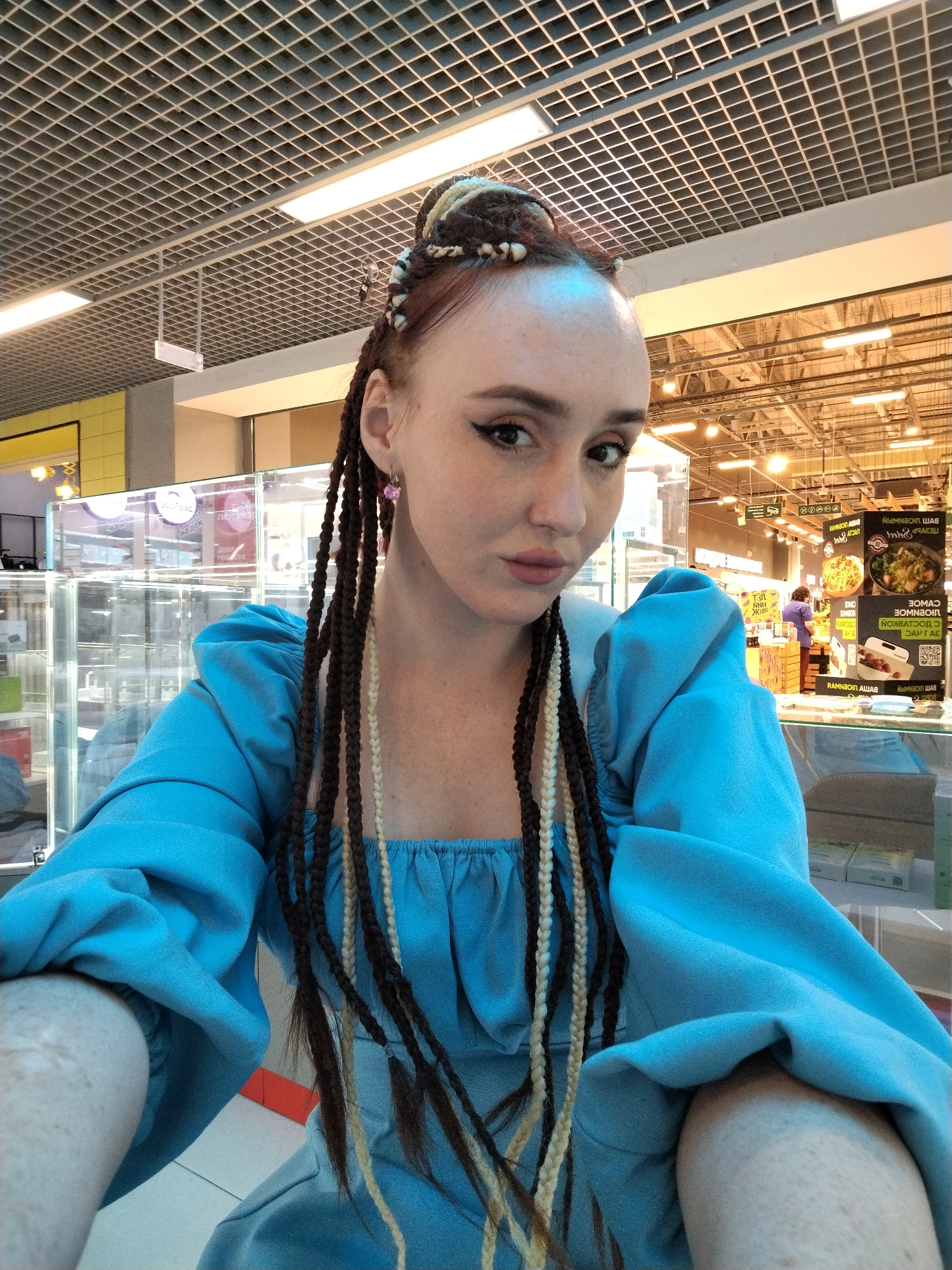 A person, identifiable as a female, is centrally positioned in a selfie-style photograph, taken indoors at what appears to be a shopping mall or retail space in Izhevsk, Russia. The person is fair-skinned with visible freckles and is wearing her hair in a distinctive style involving dark and blonde braids, some of which frame her face. She is dressed in a bright blue garment that features puffed sleeves.
In the background, out-of-focus retail displays and overhead lighting fixtures suggest the scene is a commercial environment, and Cyrillic signage is seen, confirming the location as Izhevsk, Russia. The lighting is bright and even, typical of an indoor public space. There are other individuals in the background, further indicating the public setting. The image is taken at eye level from a slightly higher vantage point, as would be expected from a selfie angle.