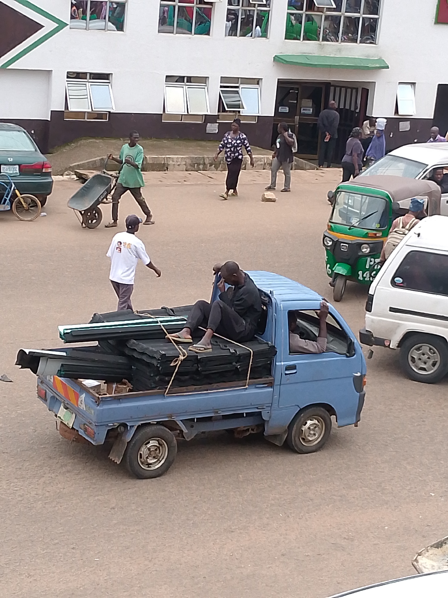 The image depicts a street scene in Bukuru, Nigeria, with a blue utility truck carrying a load of dark, corrugated roofing materials. Two individuals are visible in the truck bed; one is lying down, and another is seated. A third person, wearing a white t-shirt with images on the back, walks away from the truck. In the background, a green auto-rickshaw is parked near a white van. A man in a green shirt pushes a wheelbarrow, and other pedestrians are present near a building with windows. A dark-colored car is parked on the left side of the image. The ground surface appears to be asphalt or paved road.