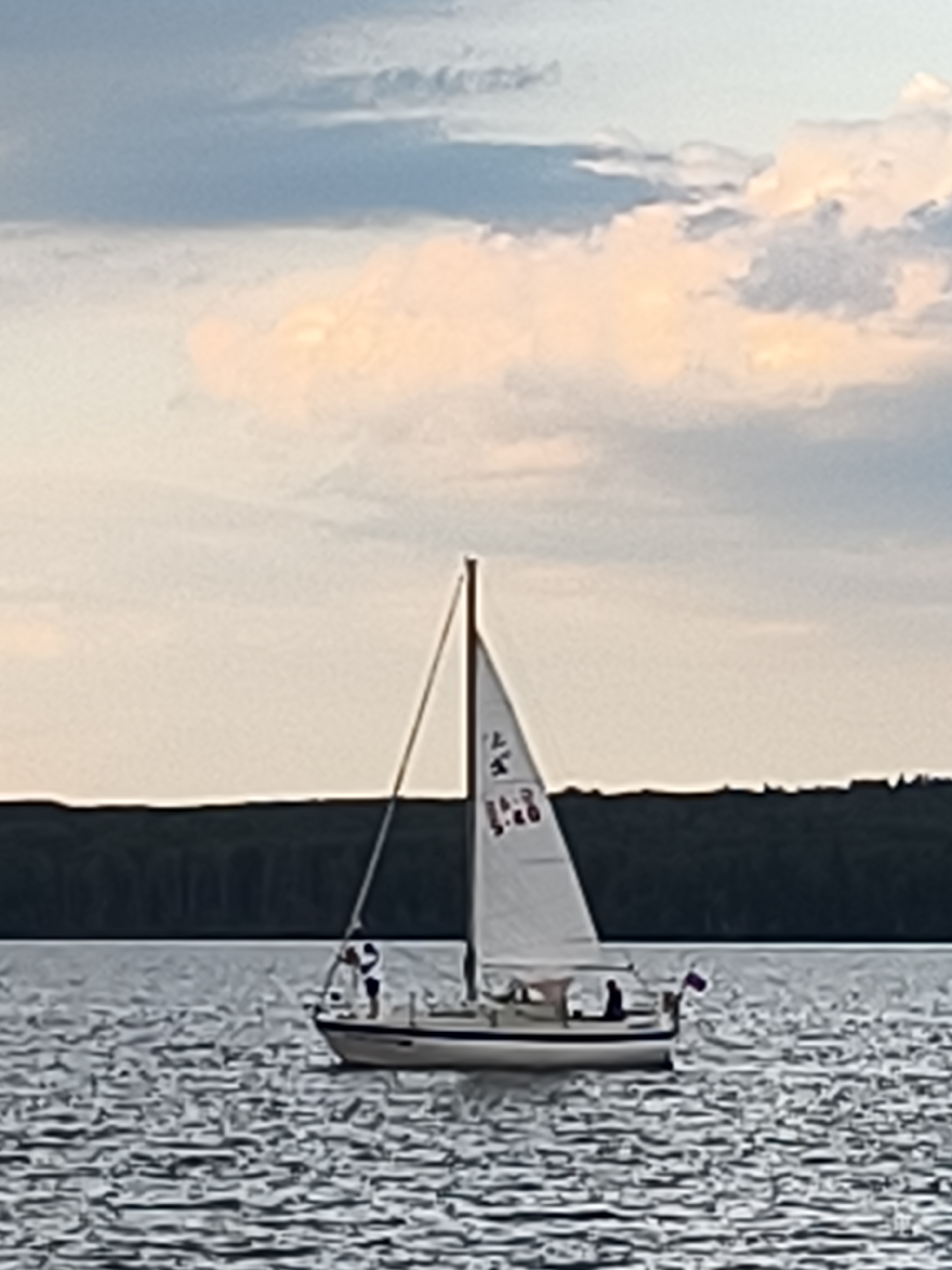 A sailboat with two people aboard sails on a body of water, likely a lake or calm sea, with a dark treeline visible in the background. The sky above is partly cloudy with muted tones of grey and orange. The sailboat is white with a dark hull and features a white sail with some lettering and numbers visible, possibly a race number or identifying mark. One person appears to be standing at the front of the boat, and another is seated or standing in the cockpit. The water is choppy with visible ripples. There is no indication of the city of Chaikovsky, Russia in the image.