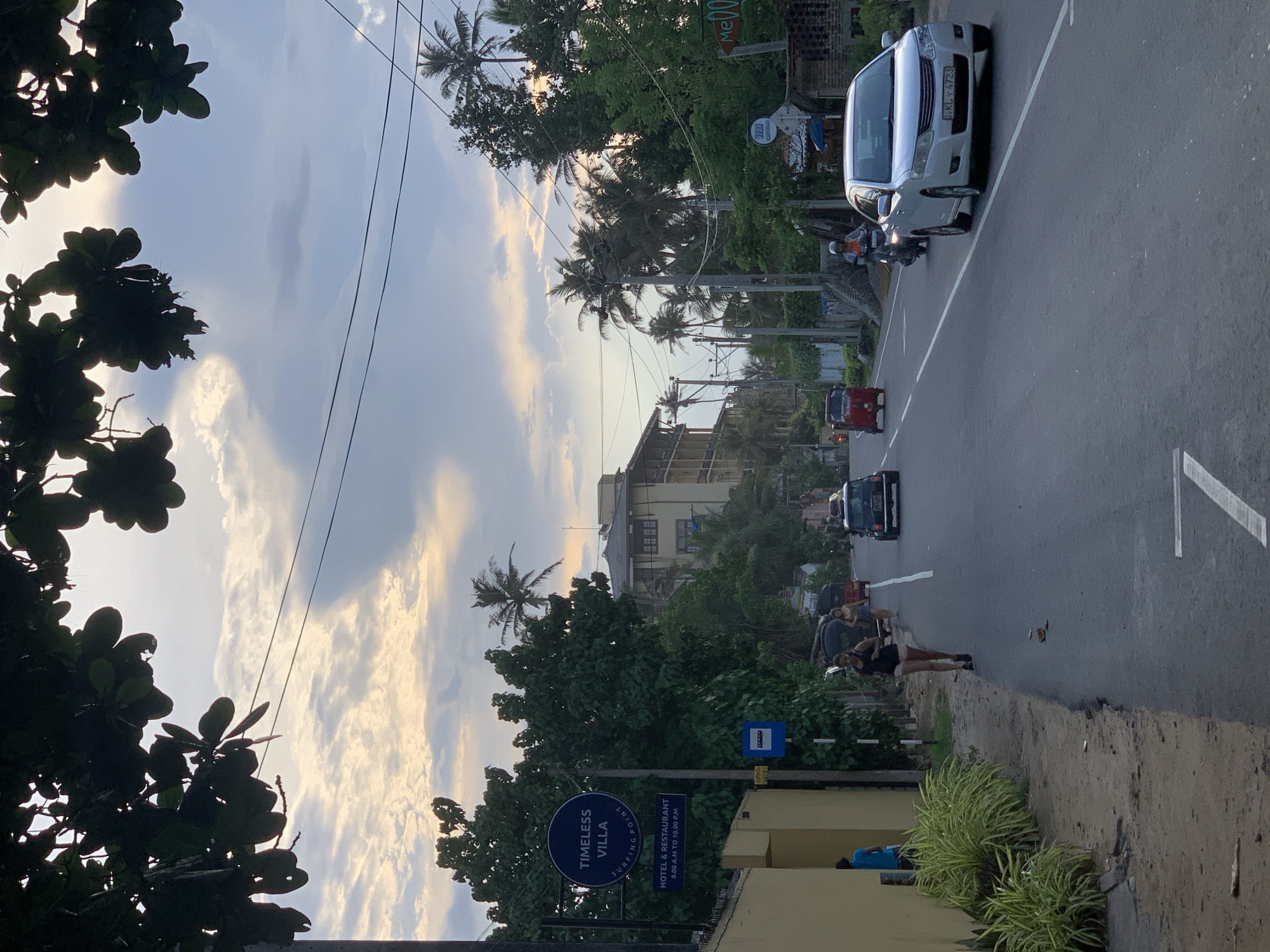 The image depicts a street scene in Ahangama, Sri Lanka. The view looks down a road with several vehicles, including a white van, a red tuk-tuk, and other cars. A couple stands on the right side of the road. Palm trees and other tropical foliage line the street. A sign for "Timeless Villa, Surfing Point Hotel & Restaurant" is visible in the foreground. Overhead power lines stretch across the sky, which is filled with clouds. The overall setting appears to be a warm, tropical climate. The license plate of the white van is KL-4723.