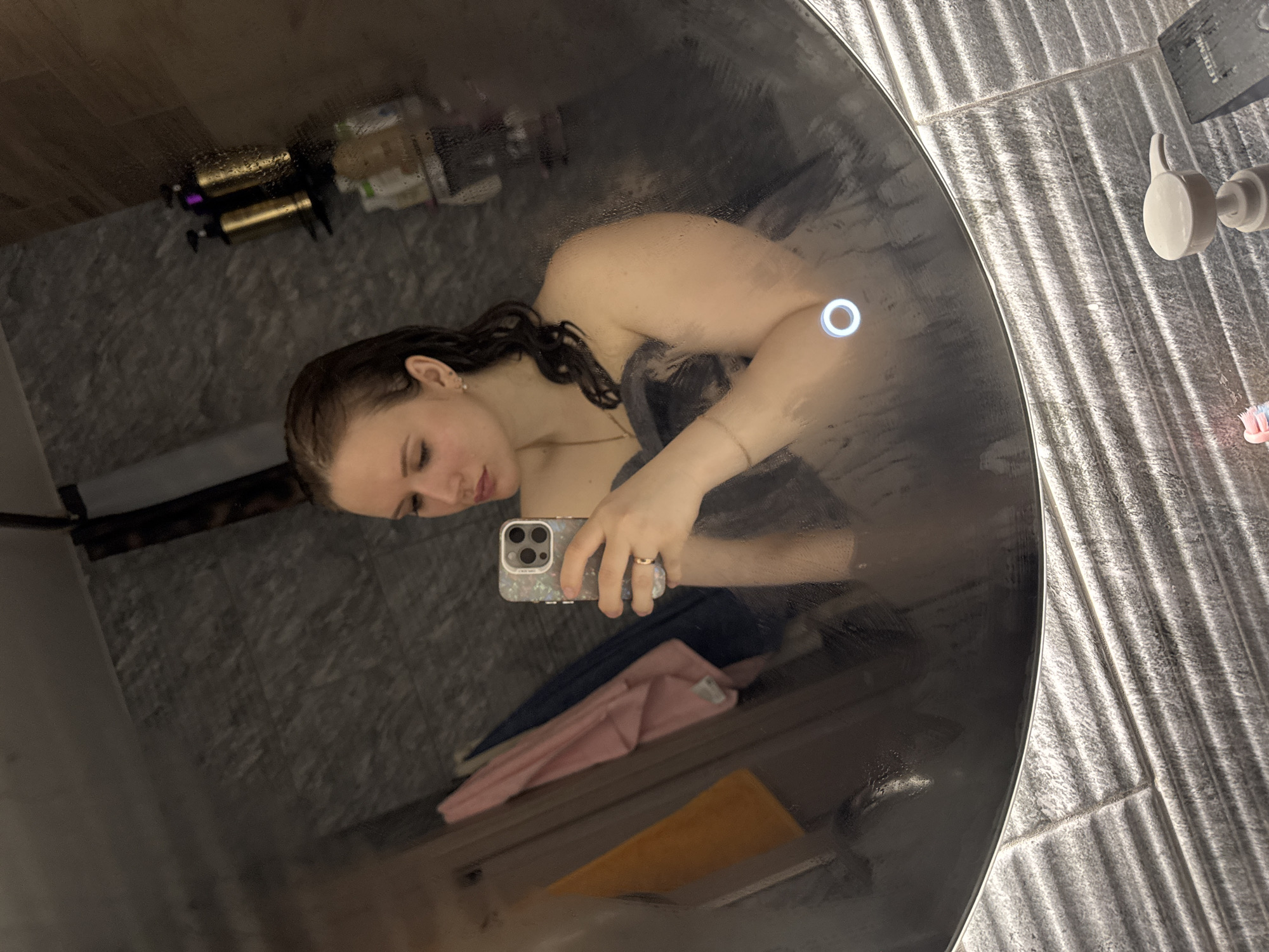 A woman is taking a selfie in a round mirror in a bathroom. She is partially wrapped in a gray towel and holding an iPhone. The mirror is foggy from moisture. There is a gray tiled wall behind the mirror, and bathroom supplies are on a shelf to the left. Two additional towels, one pink and one orange, are visible below the woman's reflection. The location is potentially in Chaikovsky, Russia, but this is not explicitly verified from the image.