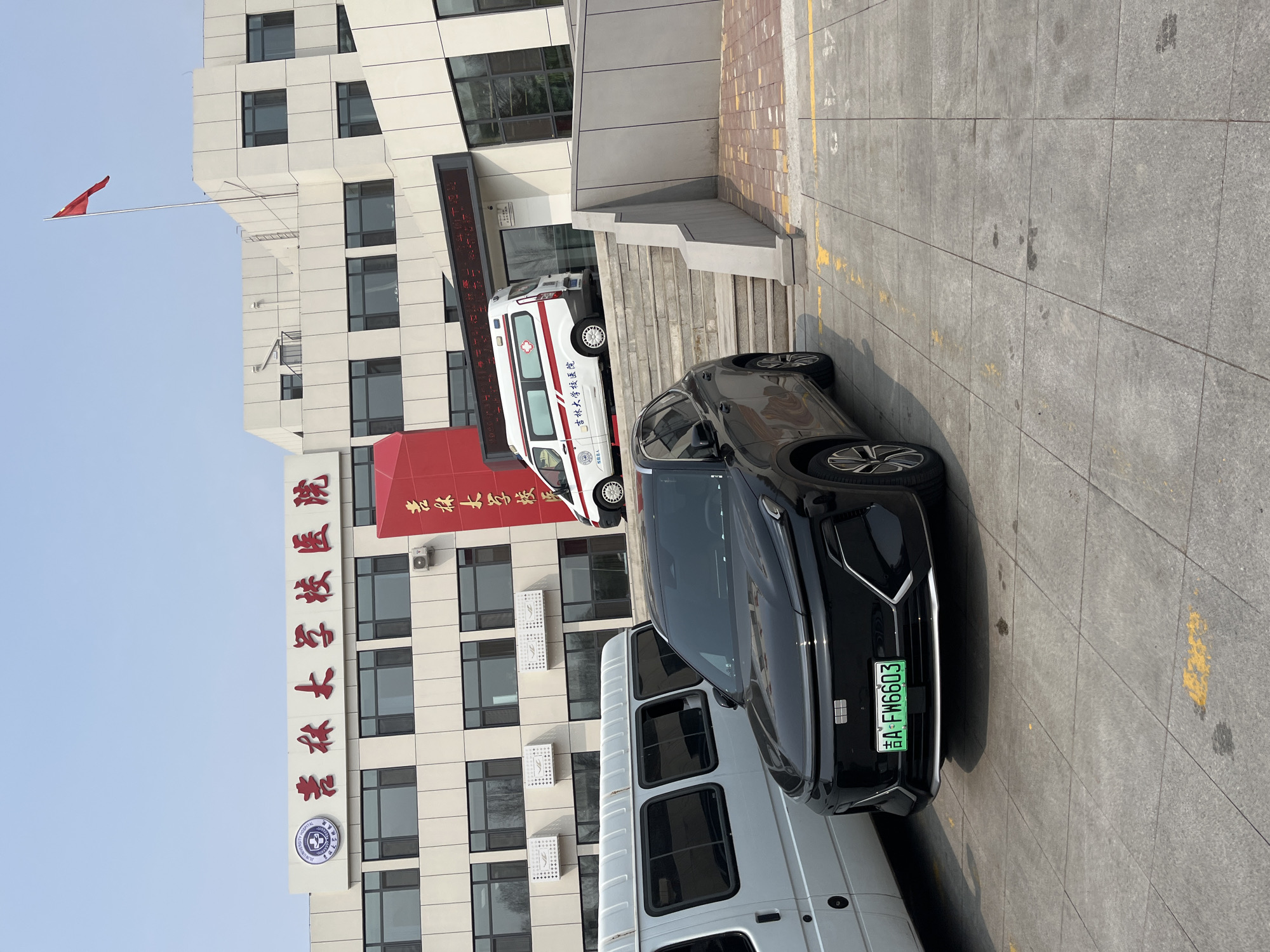 Here is a detailed description of the image:
**Scene Overview:**
The image shows a street scene, likely in Changchun City, China, in front of a building.
**Building:**
The building is a multi-story, modern structure with a white facade and dark-framed windows. A red banner or signboard is prominently displayed on the front of the building, displaying Chinese characters. A Chinese flag is visible atop the building. Based on the red text and a circular badge on the building, it appears to be the hospital of Jilin University ("吉林大学校医院").
**Vehicles:**
Several vehicles are present in the image:
* A black car is parked prominently in the foreground. It has a green license plate with the registration "吉A·FW6603".
* A white van is parked adjacent to the black car.
* A white ambulance with red markings and "吉林大学校医院" written on it is parked near the building's entrance.
**Environment:**
The scene appears to be an outdoor area in front of the building, possibly a parking area or a drop-off zone. The ground is paved with gray tiles. Yellow lines are painted on the ground, presumably to mark parking spaces.
**People and Interactions:**
There are no people visible in the frame. There is no evidence of any action.