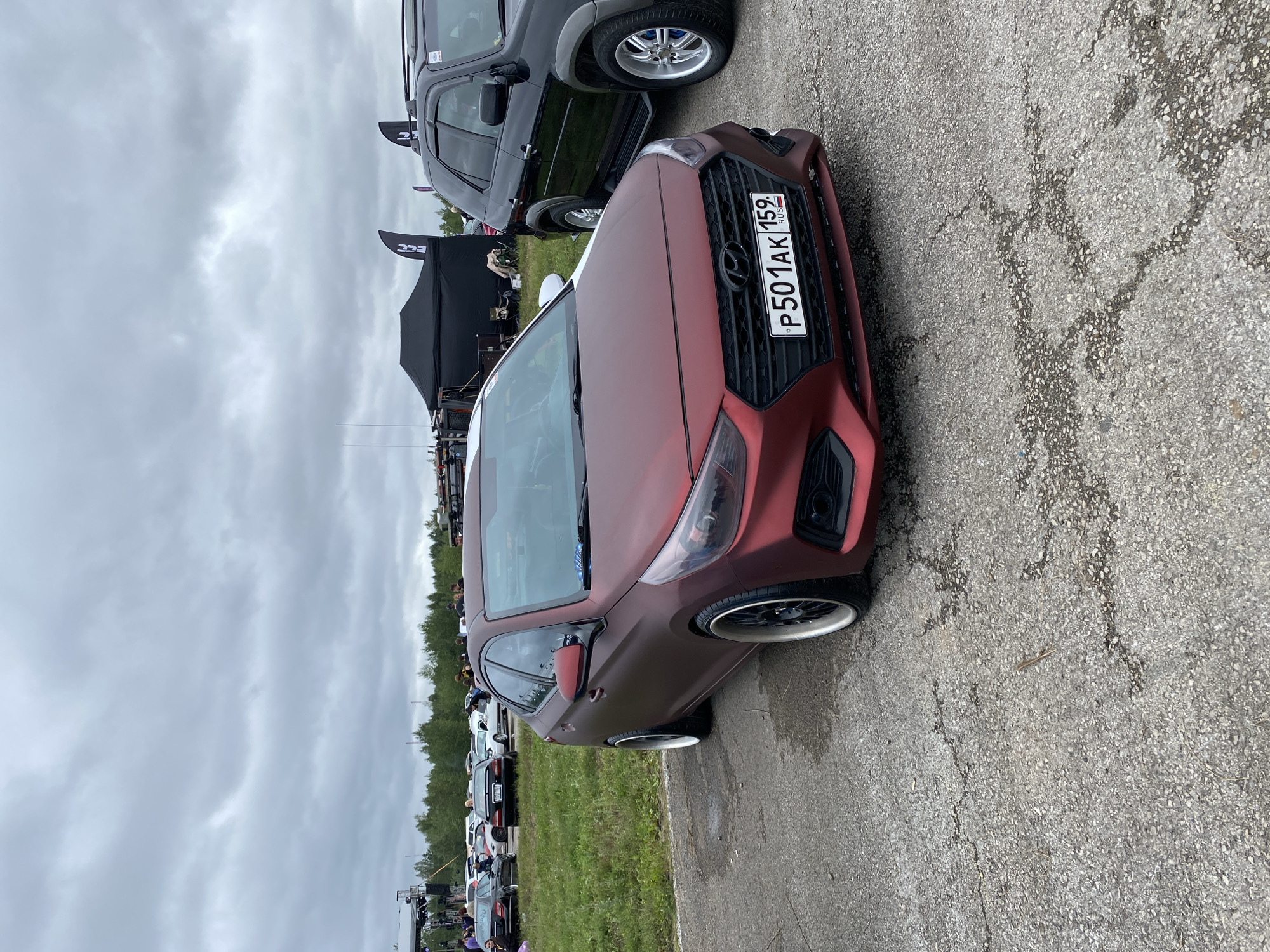 A matte red Hyundai sedan with a custom body kit and black rims is parked on a cracked asphalt surface. The car's license plate reads "P501AK 159 RUS". In the background, there are other vehicles, a black tent with banners, and a crowd of people. The sky is overcast with dark clouds. The scene appears to be an outdoor event or car show. The presence of a Russian license plate indicates the location is in Russia. The specific mention of Perm, Russia, cannot be confirmed from the visual information alone.