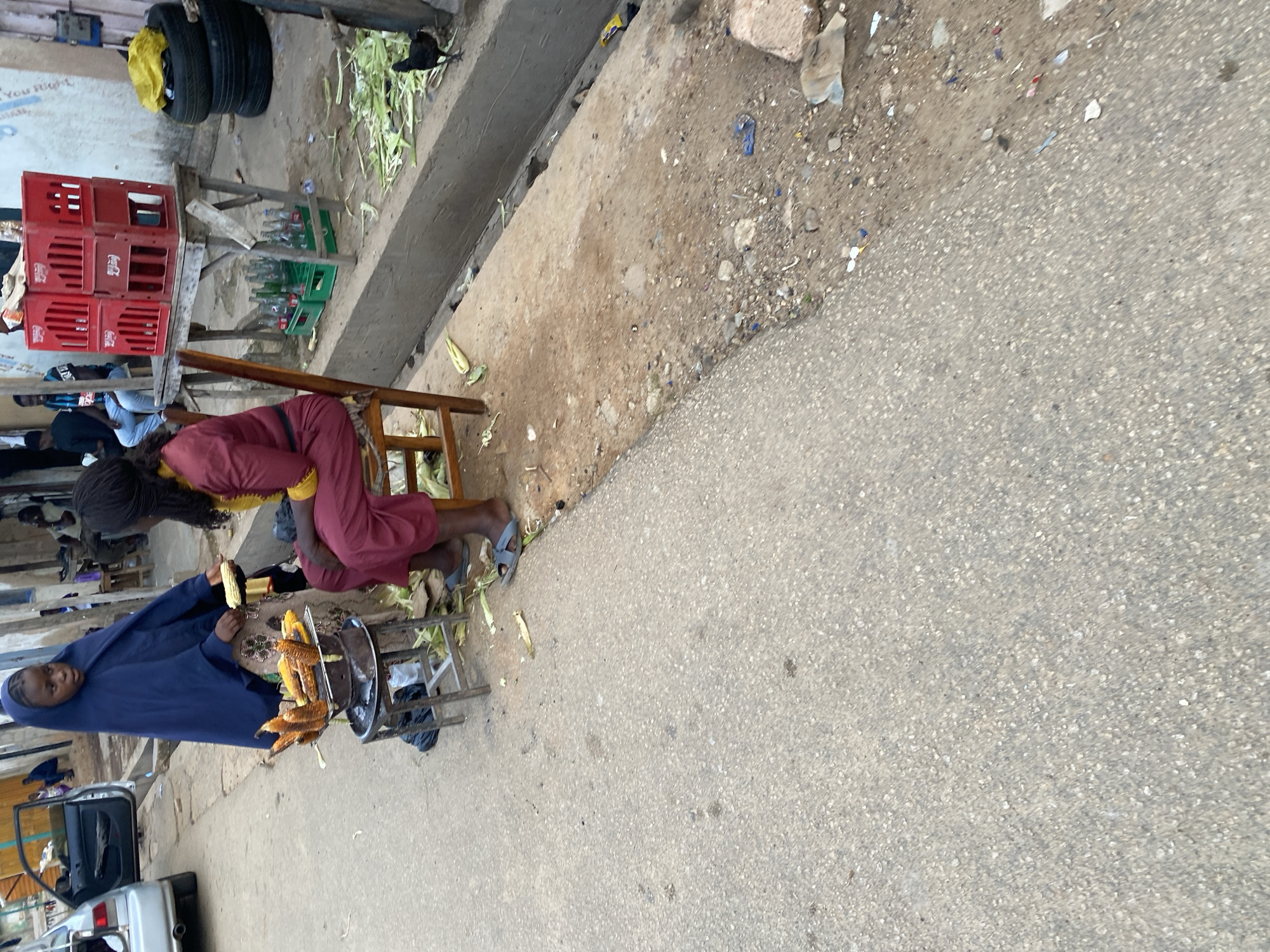 This media file captures a street scene in Jos, Nigeria, likely during daytime with good weather. The focus is on a woman selling roasted corn from a cart. She is dressed in a blue abaya and appears to be interacting with a customer who is wearing a red dress and yellow top, holding a piece of corn.
In the background, several stacked red Coca-Cola crates are visible, along with green plastic crates that might contain drinks. There are also discarded corn husks and other debris scattered on the ground, indicating a busy market or roadside vending area. The ground is a mix of dirt and asphalt. A car is parked on the left side of the frame, partially obscuring some of the vending setup.
The overall impression is that of a typical roadside market scene, highlighting daily commerce. The faces of the subjects are not clearly visible, making it difficult to discern specific emotions, but the presence of a customer and vendor suggests an ongoing transaction. The lighting appears to be natural, consistent with daylight. There is no visible text in the image.