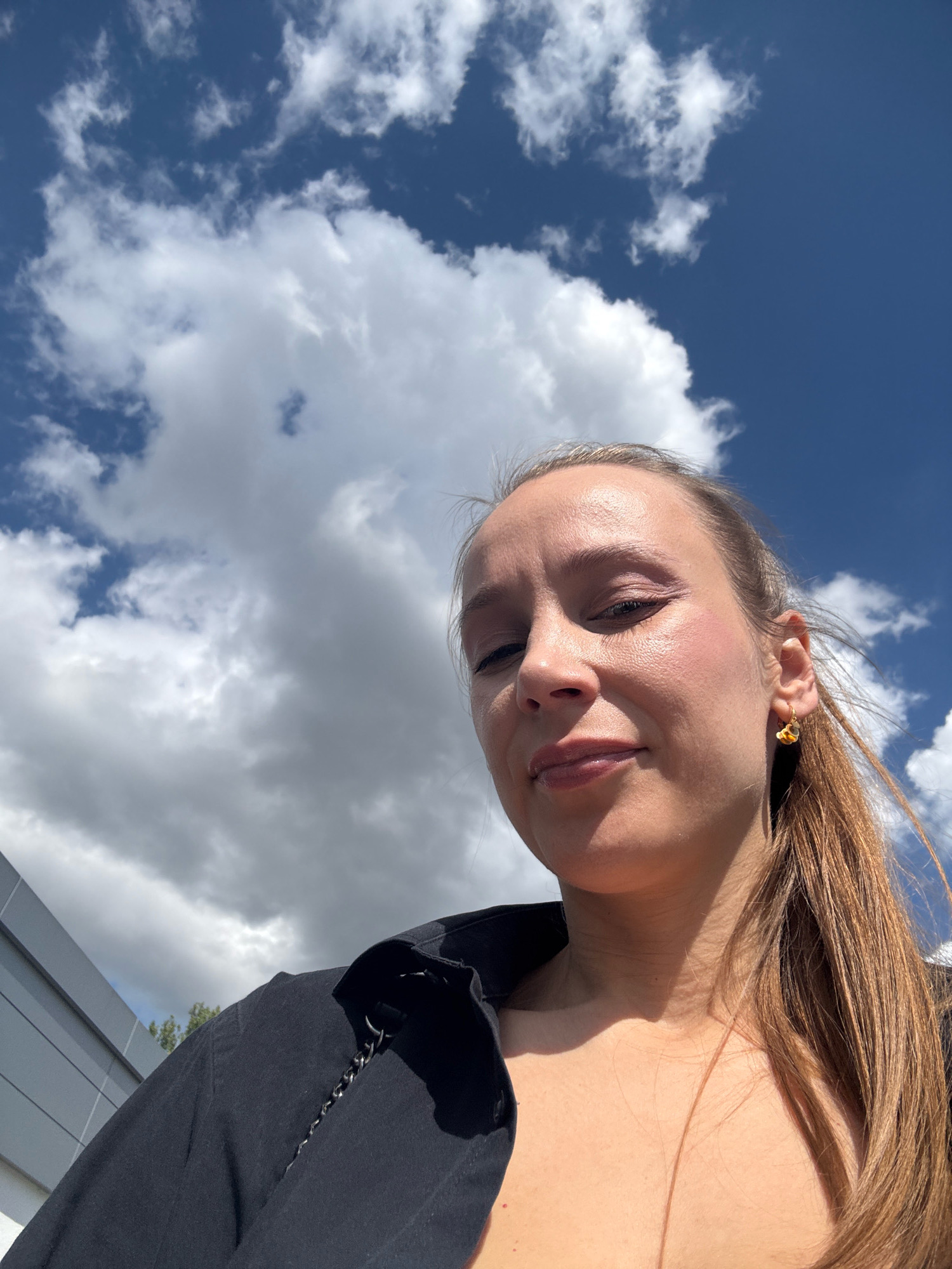 A woman is captured in a low-angle, outdoor selfie against a backdrop of a bright blue sky and white clouds. She has light brown hair pulled back into a ponytail and is wearing a black jacket. She has on makeup, including blush and eyeshadow, and is wearing a gold earring. The image appears to be taken in Überherrn, Germany, based on the location tag. The environment includes a grey building to the left, possibly indicating an urban or suburban setting. The lighting suggests it is daytime.