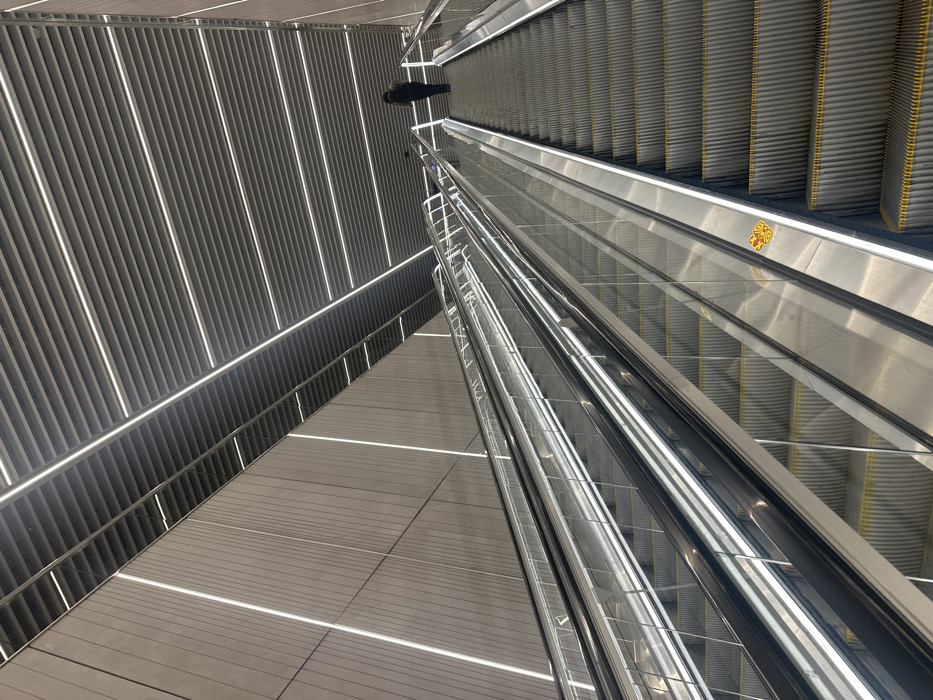 An upward view showcases the architectural design of an escalator area. The escalator, primarily silver with yellow accents, contrasts with a gray, textured ceiling and wall arrangement. Lighting fixtures create bright, linear patterns across the surfaces, emphasizing the geometric forms. A dark, silhouetted figure is briefly visible above the escalator.