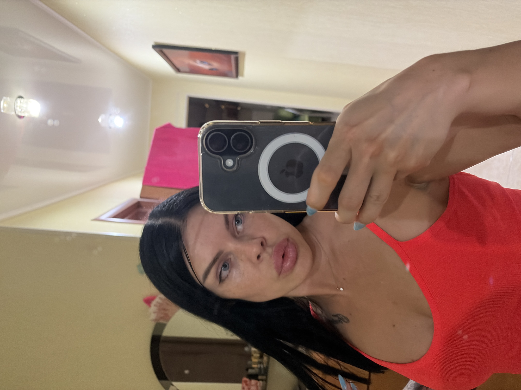 A woman with long, dark hair and blue eyes takes a selfie in a mirror. She is wearing a bright red, ribbed tank top and has light blue nail polish. A delicate necklace with a small pendant is visible around her neck. Her phone, with a black case featuring a circular white magnetic mount, is held up to take the photo. The background appears to be an indoor hallway with light yellow walls, a white, reflective ceiling with two lights, and a framed picture on the wall. A partially visible mirror to the left shows a reflection of a vanity area with various items on it and a pink towel draped over something. The scene suggests a domestic setting, possibly a home or hotel room, within the city of Ufa, Russia.