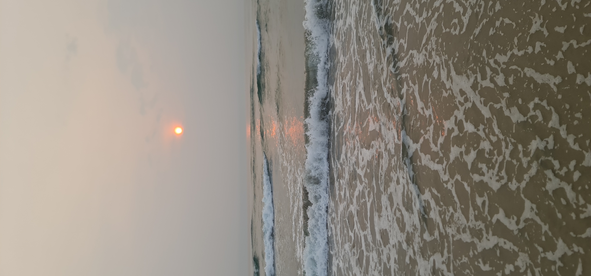 The image displays a serene coastal scene, likely captured during either sunrise or sunset in Đà Nẵng, Vietnam. The sky occupies the upper portion of the frame (when viewed correctly, as the original image is rotated). It appears hazy or lightly clouded, with a soft, diffused light quality.
A distinct, round orange sun is visible low in the sky, positioned towards the upper left. Its warm glow creates a subtle, elongated reflection on the water's surface, extending horizontally from the sun across the ocean.
The lower and right-hand side of the image features the ocean and its shoreline. Gentle waves are seen breaking and receding, creating dynamic patterns of white sea foam over what appears to be wet sand or shallow water. The water itself has a muted, sandy-brown hue, interspersed with the bright white of the foam.
There are no people, animals, or man-made objects visible, emphasizing the natural beauty and tranquility of the beach environment. The overall mood is peaceful and calm, characterized by the soft light and rhythmic movement of the waves. No visible text is present in the image.