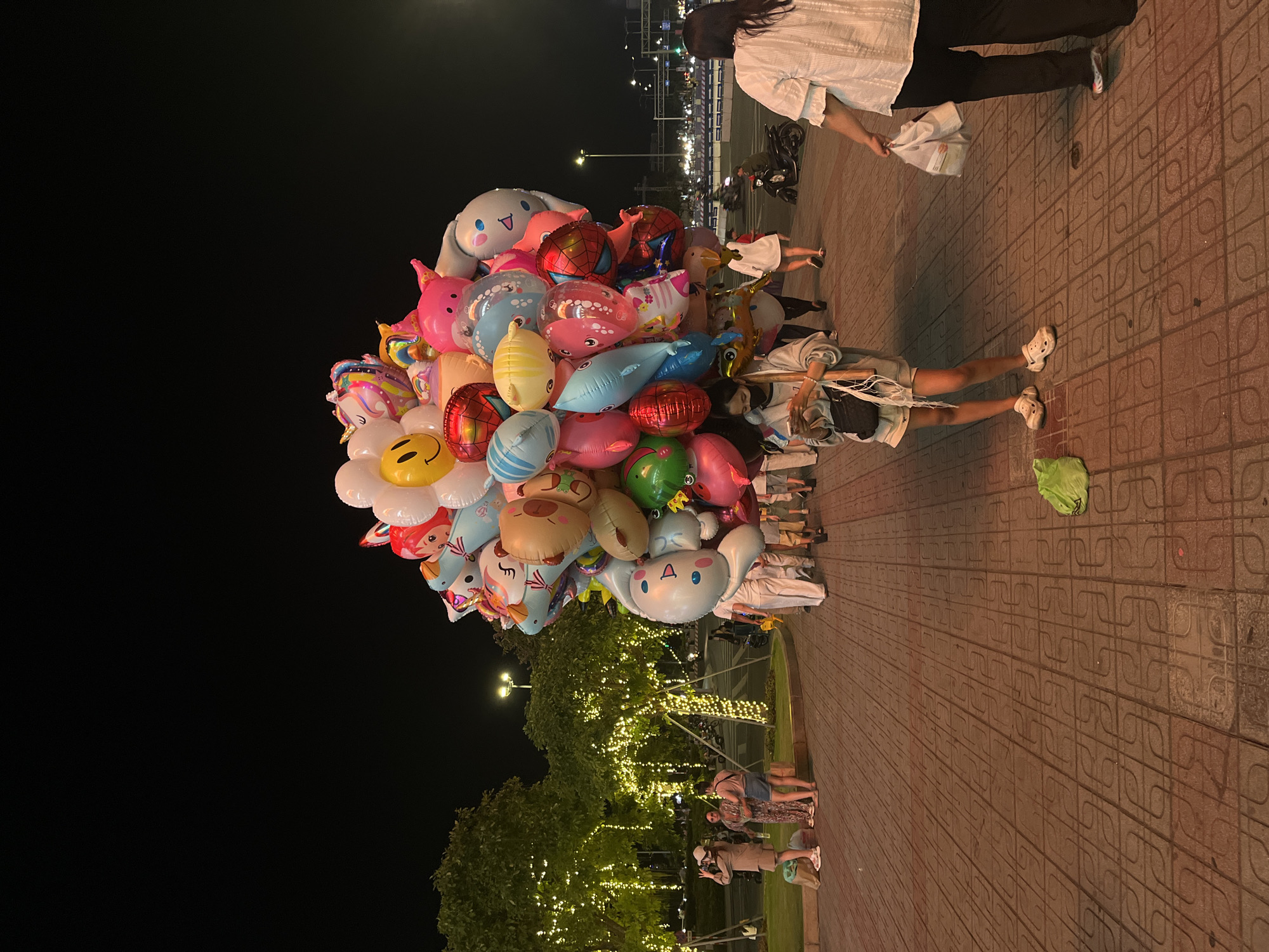 Captured at night on a brick-paved pedestrian area in what is likely the city of Đà Nẵng, Vietnam, a balloon vendor stands holding a large cluster of colorful helium balloons of varying shapes and designs, including a smiling daisy, a unicorn, a dolphin, and several cartoon characters. The vendor is standing next to a woman. The background includes city lights and trees strung with decorative lights. Other people are also in the scene.