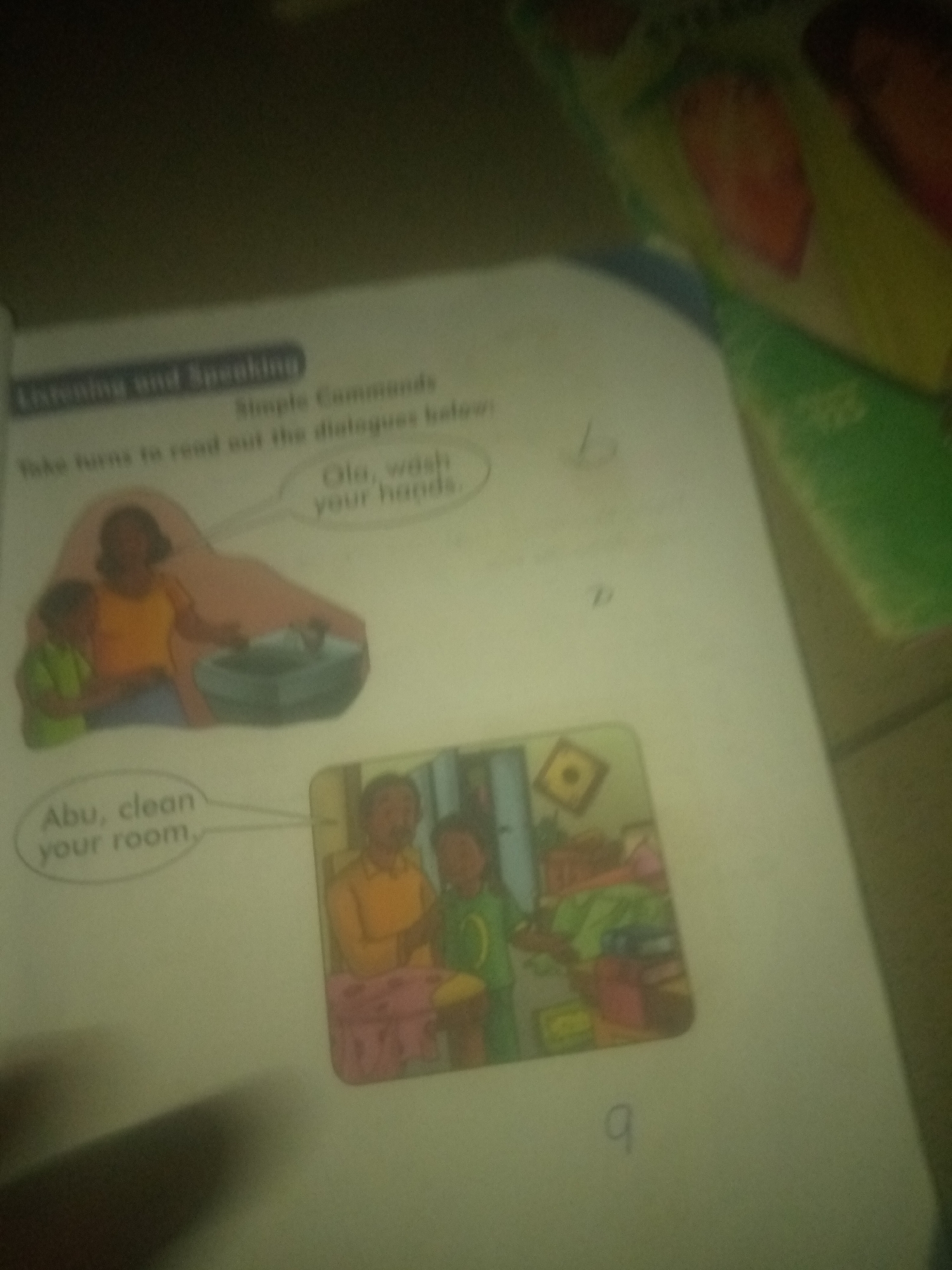 The image displays a page from a children's learning material, possibly a book or worksheet, with text and illustrations. The text at the top reads "Listening and Speaking" and "Simple Commands." Below this, it instructs: "Take turns to read out the dialogues below."
One dialogue bubble, associated with an illustration of a woman and a child by a sink, reads "Ola, wash your hands." The illustration depicts the woman gesturing towards the sink, with the child standing beside her.
Another dialogue bubble, associated with a separate illustration, states "Abu, clean your room." This illustration shows a man and a child in a room that appears to be cluttered.
The environment is a flat surface, likely a table, with the book or worksheet placed on it. In the background, out of focus, there are elements that appear to be other books or learning materials with colorful images of fruits and green surfaces. The lighting is dim, creating shadows. The overall impression is that of a learning activity in progress.
There is no information in the image that can be used to determine its location in Jalingo, Nigeria.