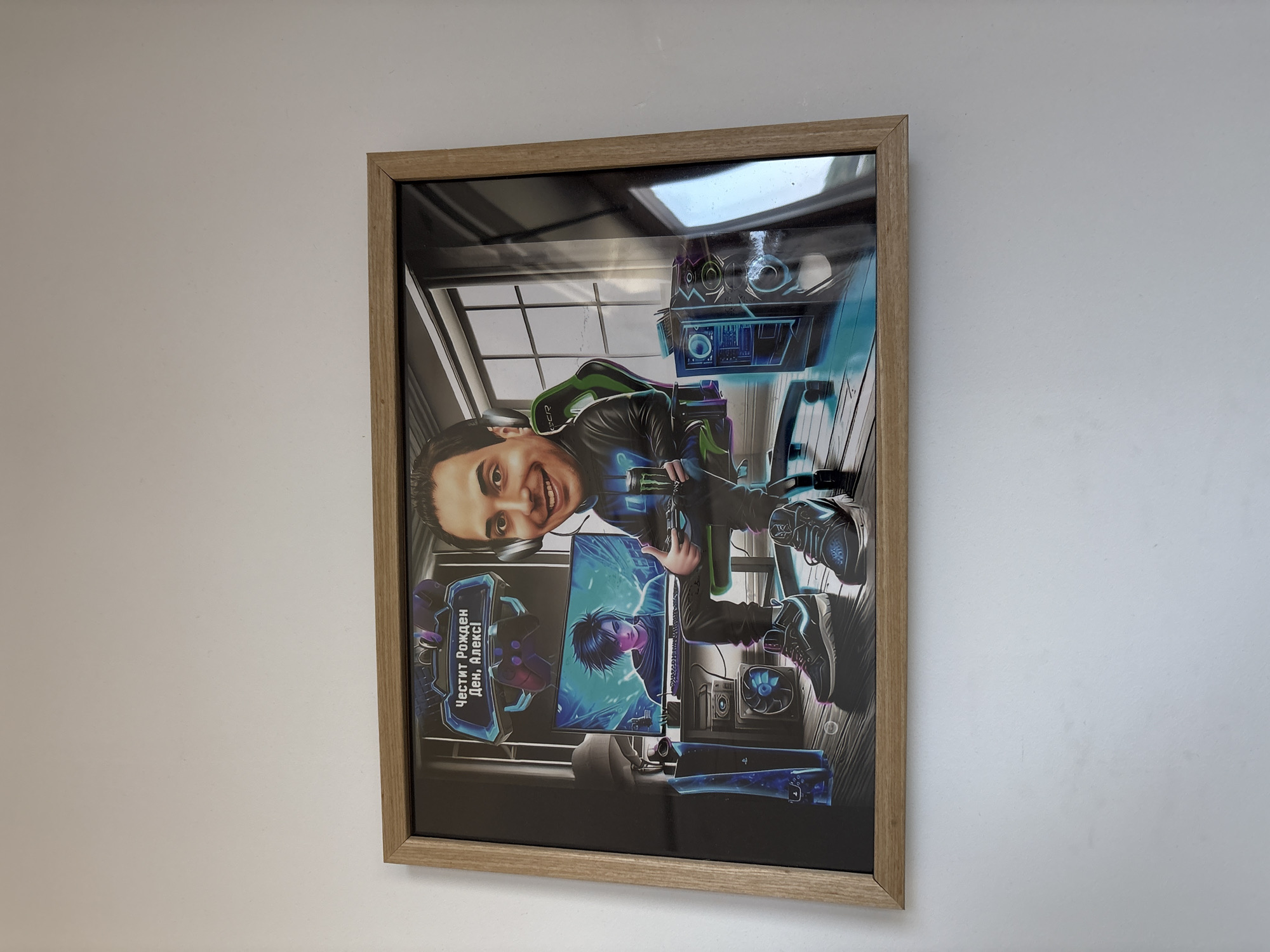 A framed picture hangs on a white wall. The picture is a cartoon-style depiction of a man sitting in a gaming chair, holding a controller. He is positioned in front of a gaming setup, including a computer monitor displaying an anime-style image and other gaming equipment. A sign above the monitor is written in Cyrillic script. The setting appears to be a gaming room. There is no indication this image is in Lovech, Bulgaria.