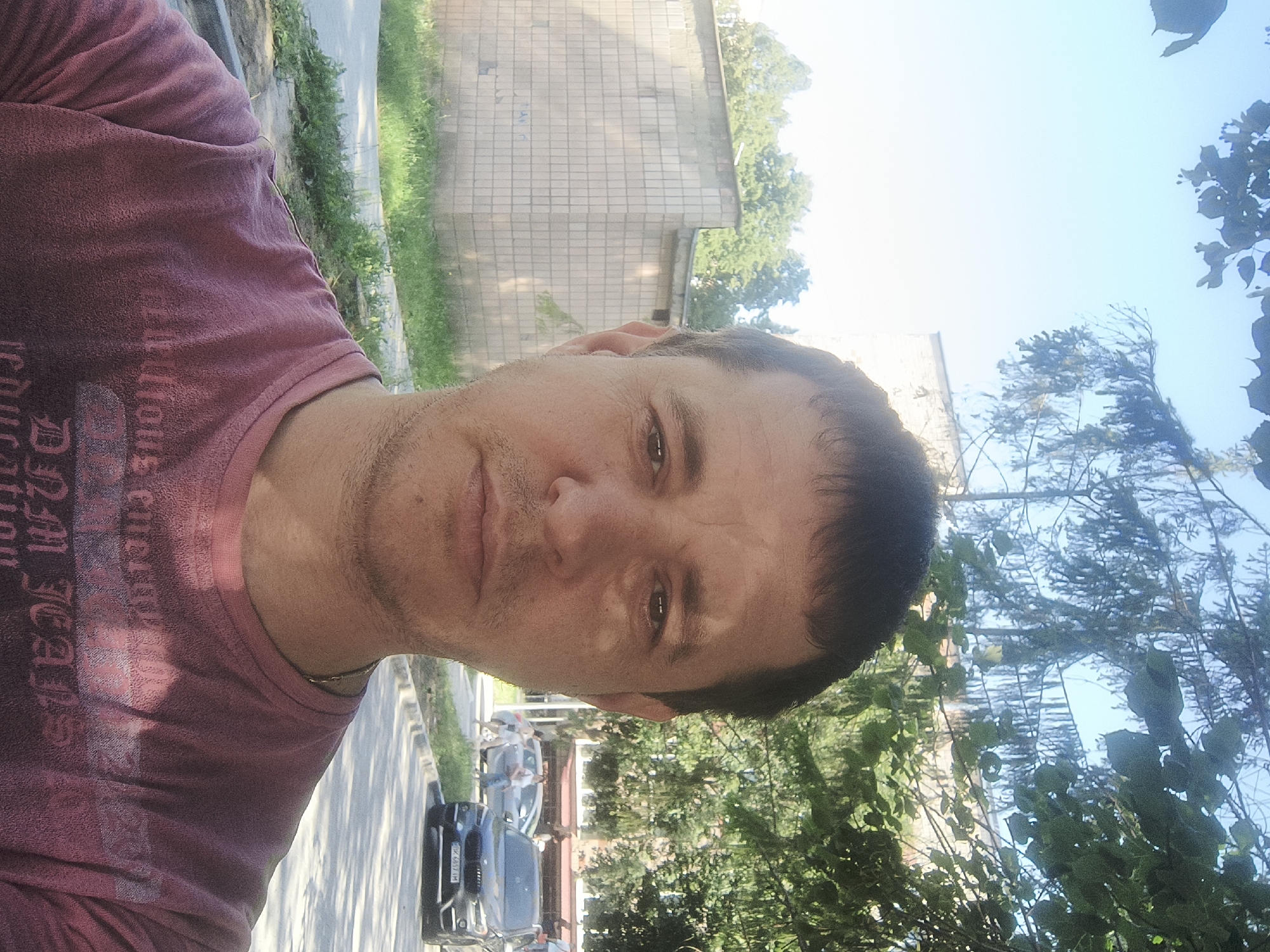 A medium shot selfie captures a Caucasian male with short, dark hair and a light stubble, looking directly at the camera. He is wearing a faded pink t-shirt with a dark graphic design on the front. The man is positioned in the foreground, with his face and upper torso filling most of the frame.
The background indicates an outdoor urban setting. To the left, a pale brick building with a tiled facade is visible, surrounded by overgrown grass and a sidewalk. Further back, trees with green foliage are present, and a section of a building with multiple windows can be seen behind them. To the right, a paved road runs horizontally. On the road, a dark-colored SUV is parked, and a silver sedan is driving away. Two individuals, a female and a male, are walking on the sidewalk in the distance, near the silver car. The sky above is a clear, bright blue. The overall lighting suggests a sunny day. The location is identified as Rivne, Ukraine.