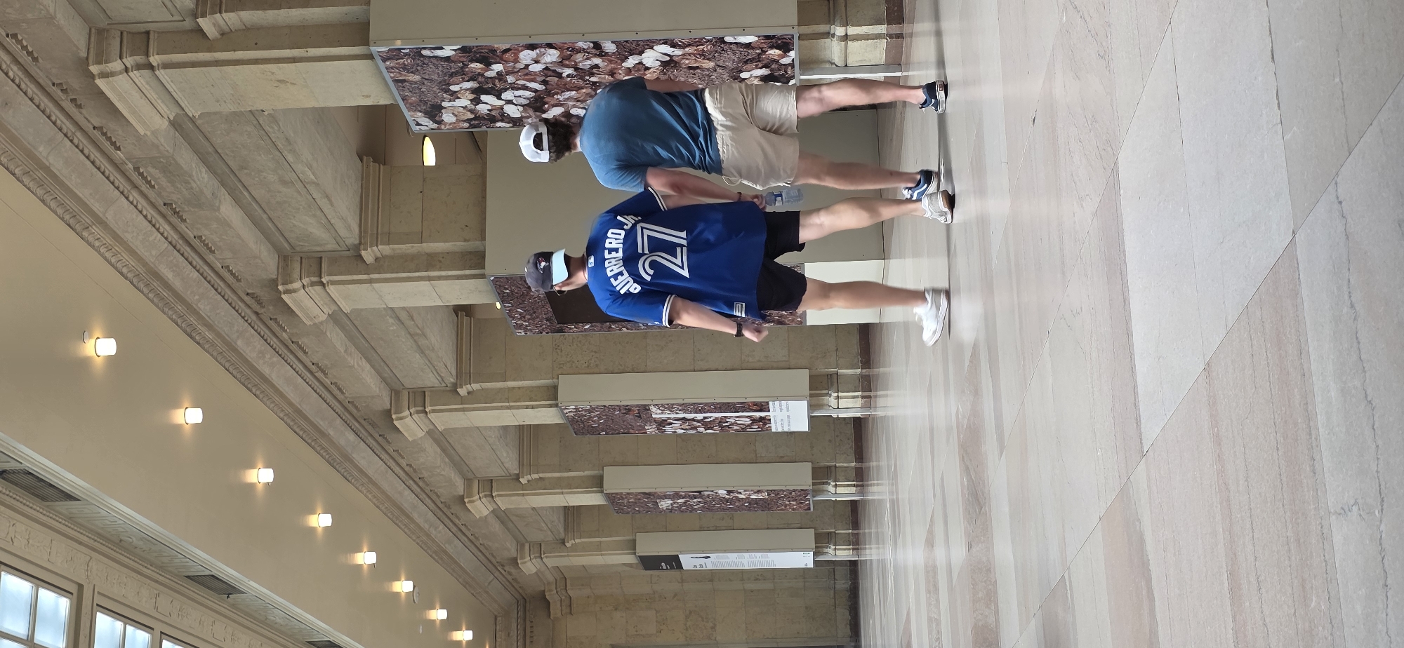 Two individuals, identified as males by their attire, walk away from the camera in an interior hallway of a large building. The person on the left wears a blue baseball jersey with "GUERRERO JR" and the number "27" in white lettering, black shorts, a dark baseball cap, and white sneakers. The person on the right wears a teal t-shirt, light-colored shorts, and dark sneakers.
The hallway features a high, arched ceiling with multiple recessed lights. The walls are constructed of light-colored stone, with ornate decorative molding running along the top. Several large display panels, featuring a close-up, abstract pattern of natural elements like leaves and nuts, are positioned along the right wall. The floor is a polished stone tile in a light, neutral color with subtle veining. The overall impression is that of a museum or a grand public building.
The image is likely taken within the Royal Ontario Museum (ROM) in Toronto, Canada, based on the distinctive architecture and the presence of large display panels.