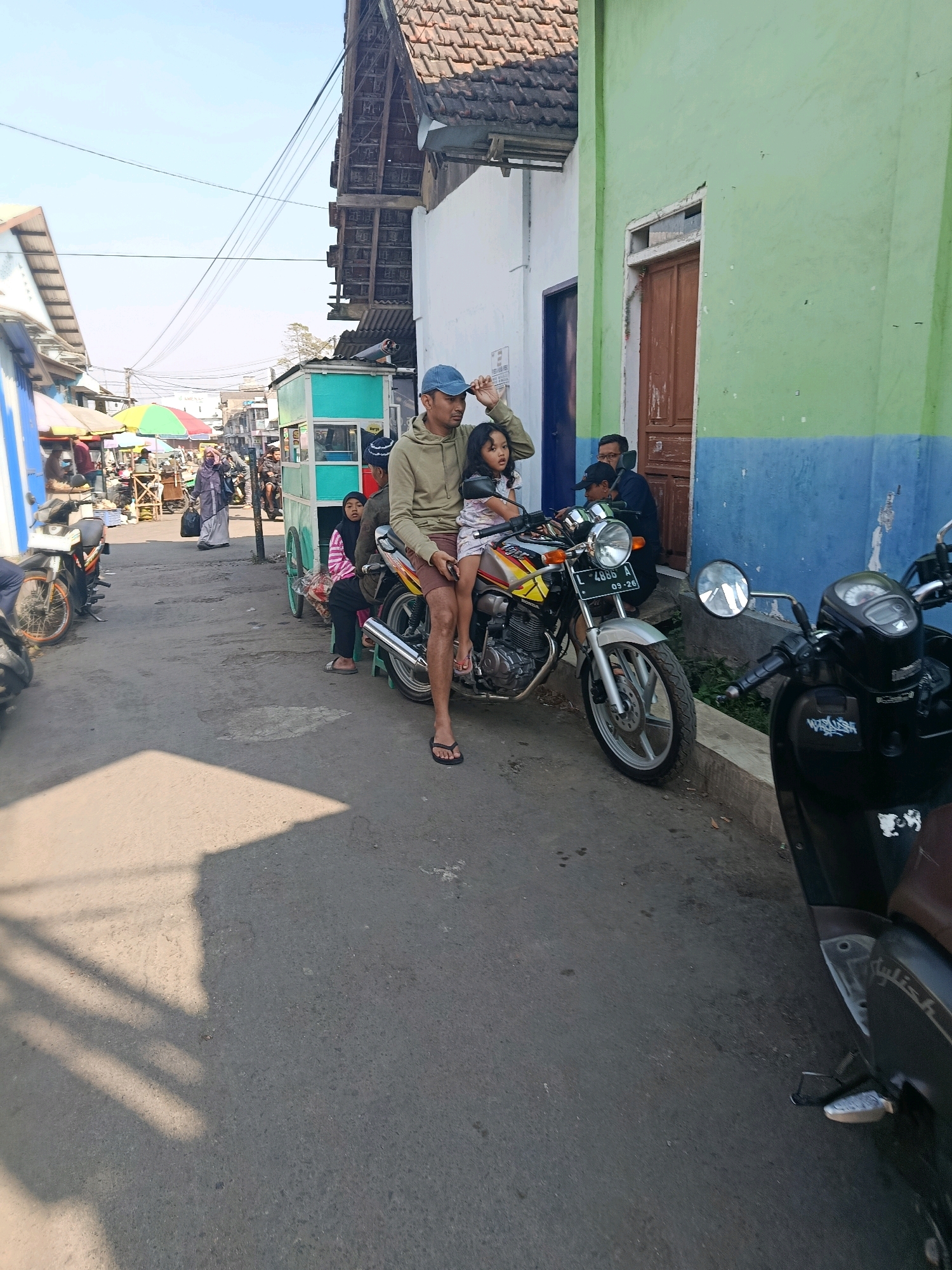 A street scene in Bululawang, Indonesia features a man wearing a light brown hooded sweatshirt and a blue baseball cap, sitting on a motorcycle with his daughter in front of him. The daughter, a young girl with long dark hair, is wearing a light-colored dress with a pattern. Behind them, a woman in a light purple hijab and long dress carries a black bag. Further back, another person in a dark blue shirt and cap is seated near a green and blue building with a wooden door. The street is paved and appears to be a market area, with stalls and colorful umbrellas visible in the background. Several other motorcycles are parked along the side of the street. The overall lighting suggests a sunny day.