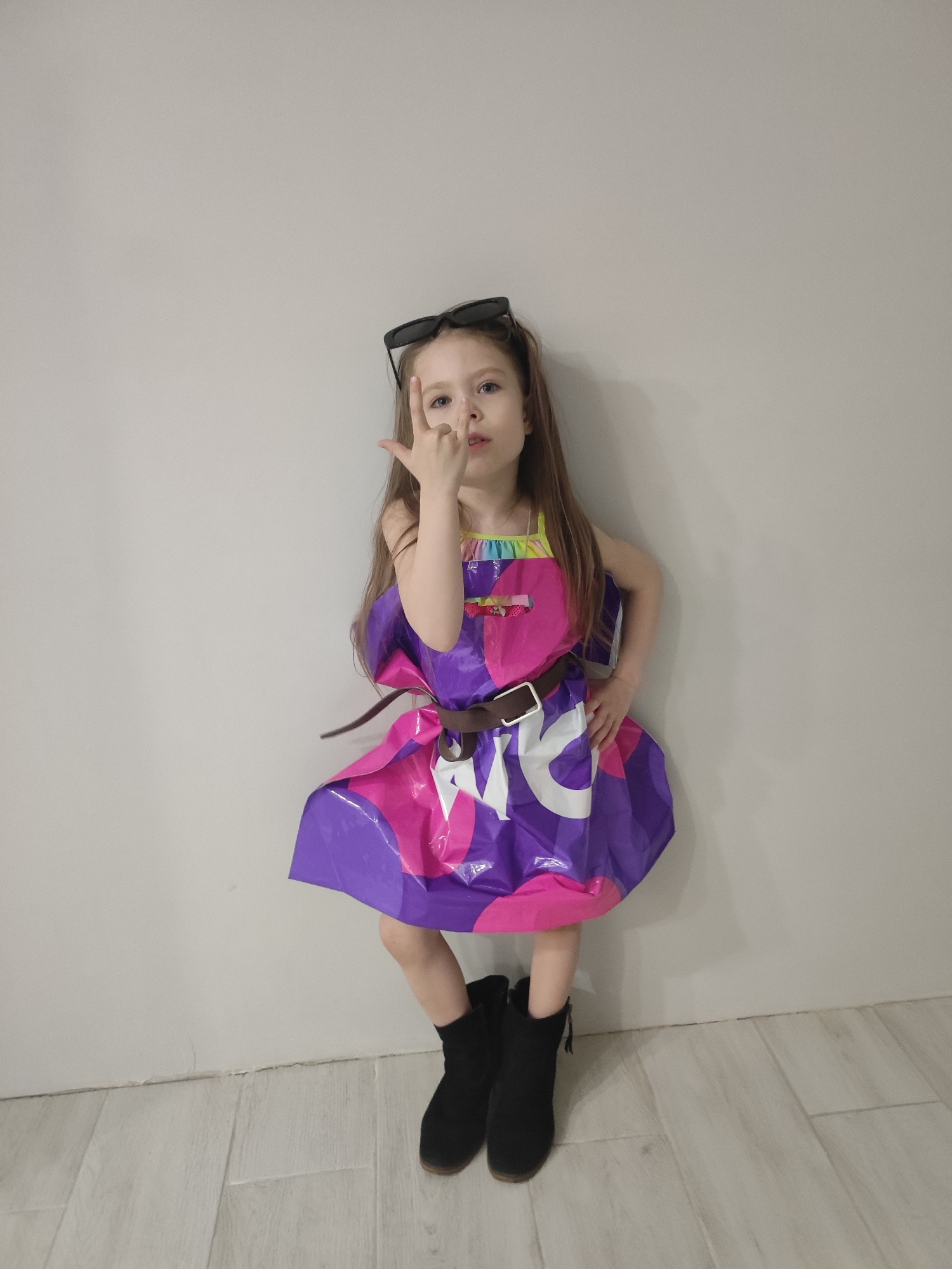 A full shot depicts a young girl standing against a light gray wall, striking a pose. She wears a dress constructed from a purple and pink plastic shopping bag, cinched at the waist with a brown belt. On top of her head are sunglasses. She is gesturing with her right hand, with her index and pinky fingers extended. She is wearing black ankle boots. The floor is made of light wood-look tiles. The picture was taken inside in Votkinsk, Russia.