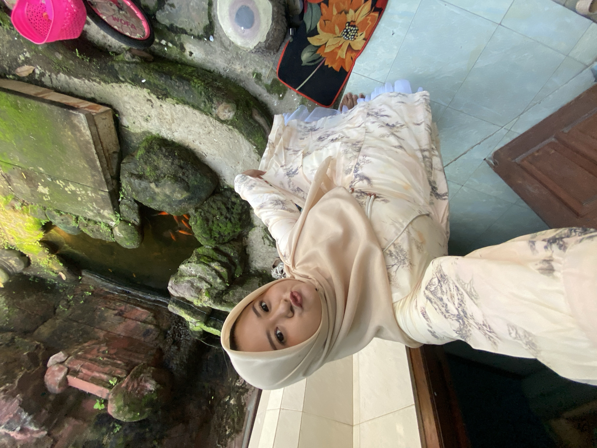 A woman in a light beige hijab and a long-sleeved, light-colored dress with a faint tree pattern is posing for a photo indoors, near an indoor fish pond. The location is in the city of Gelam, Indonesia. The pond is surrounded by moss-covered rocks, some of which have orange fish visible in the water. To the left of the pond, there is a pink plastic basket and a child's bicycle. Behind the woman, there's a black rug with a floral design. The floor is tiled with light blue tiles. To the right of the woman, there is a dark wood door.