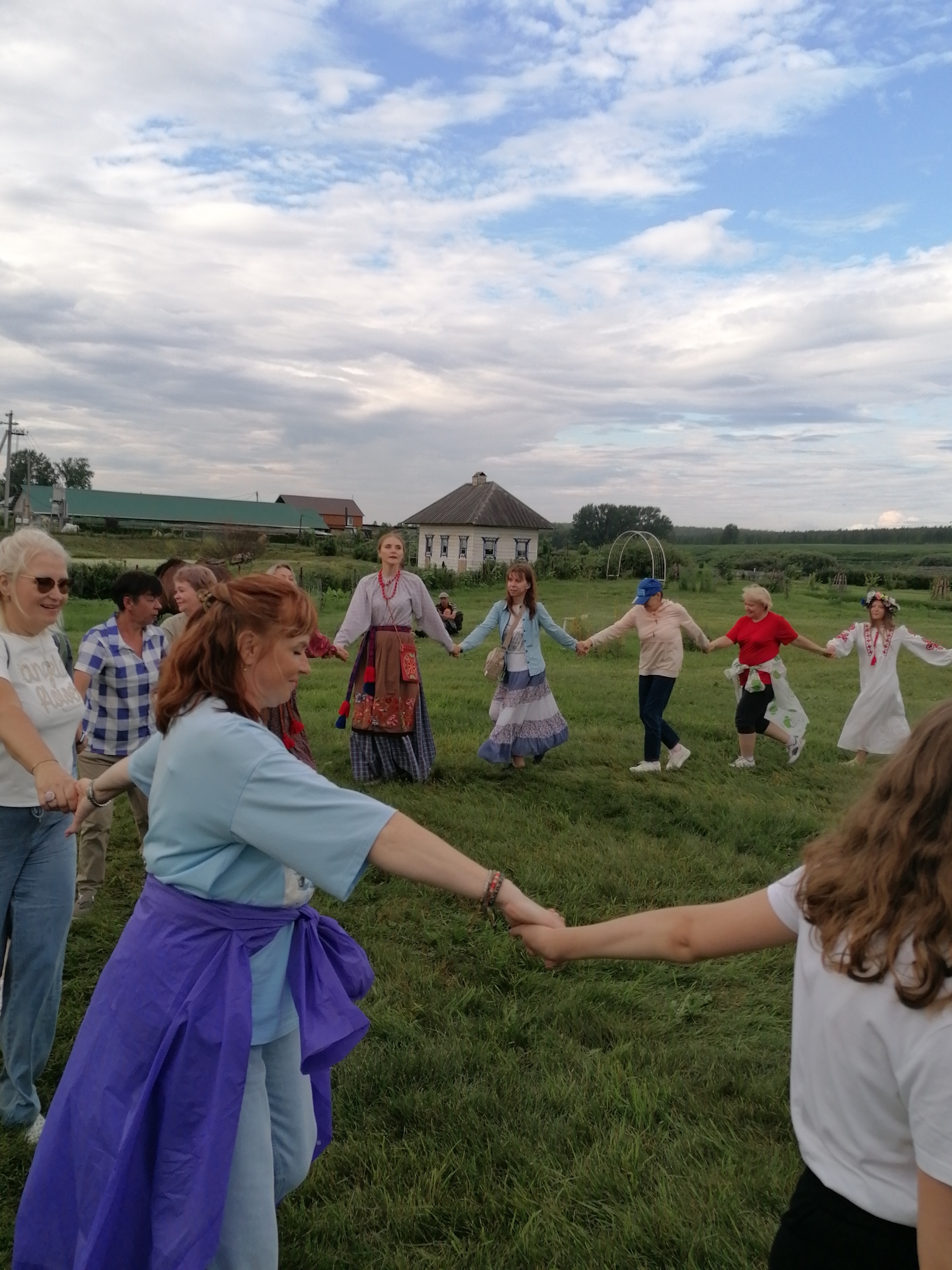 In the rural landscape near Mingali, Russia, a group of individuals is participating in a traditional folk dance. Ten people, appearing to be adults and possibly some older children, are holding hands in a circular formation. They are on a grassy field, with some agricultural buildings and a house visible in the background under a cloudy sky. The people are dressed in a mix of modern and traditional clothing. Several women are wearing long skirts and embroidered blouses, suggesting a cultural event. The dance appears to be lively, with some participants in motion. The overall atmosphere is communal and celebratory.