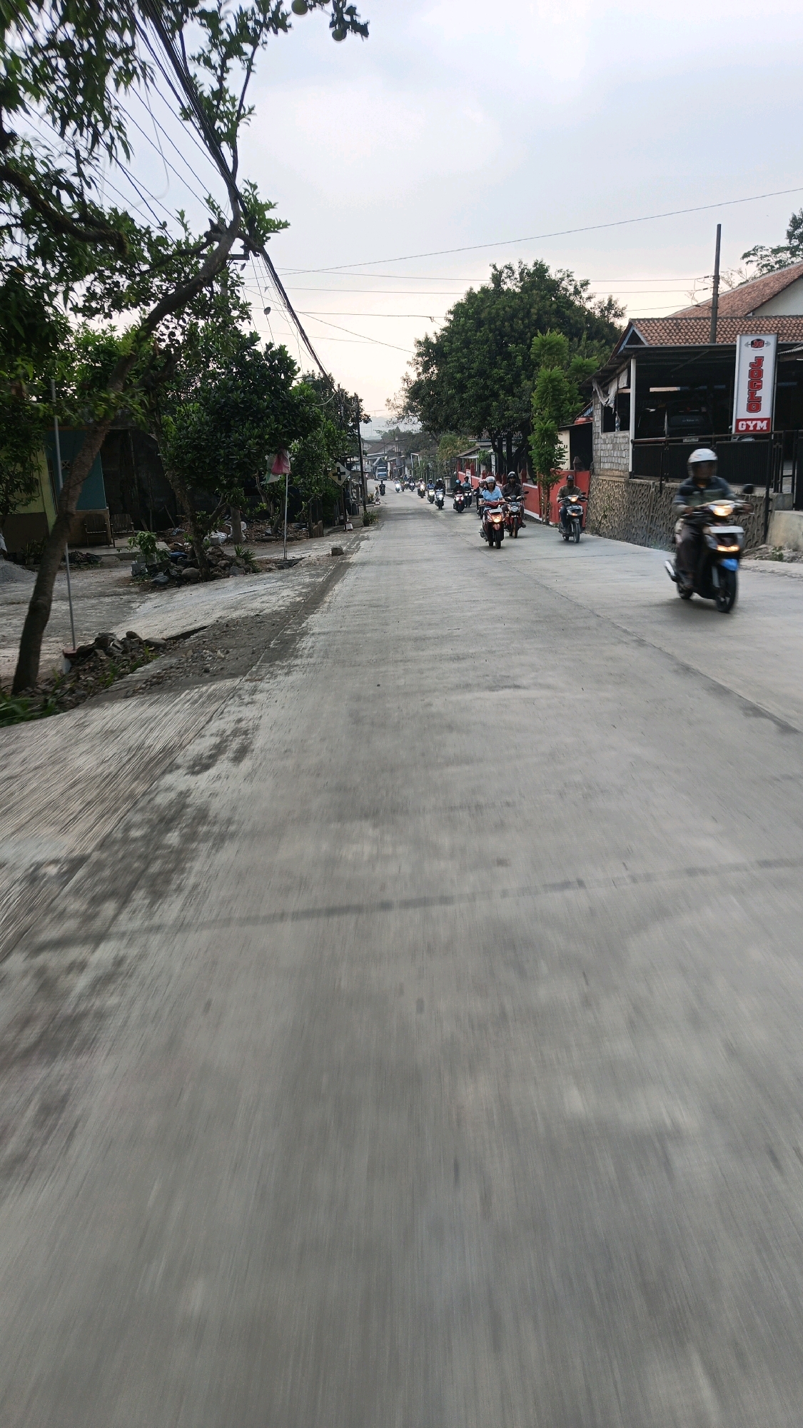 A convoy of motorcyclists is traveling down a paved road lined with trees and buildings. The road is mostly empty except for the motorcycles, which are spread out along the road. The motorcyclists are wearing helmets and casual clothing. Some of the motorcycles are red, while others are black and white. In the background, there are houses and trees. A sign with "JOGLO GYM" is visible on one of the buildings. The sky is overcast, and the sun appears to be setting or rising. The scene suggests a casual gathering of motorcyclists on a local road in Wringinputih, Indonesia.