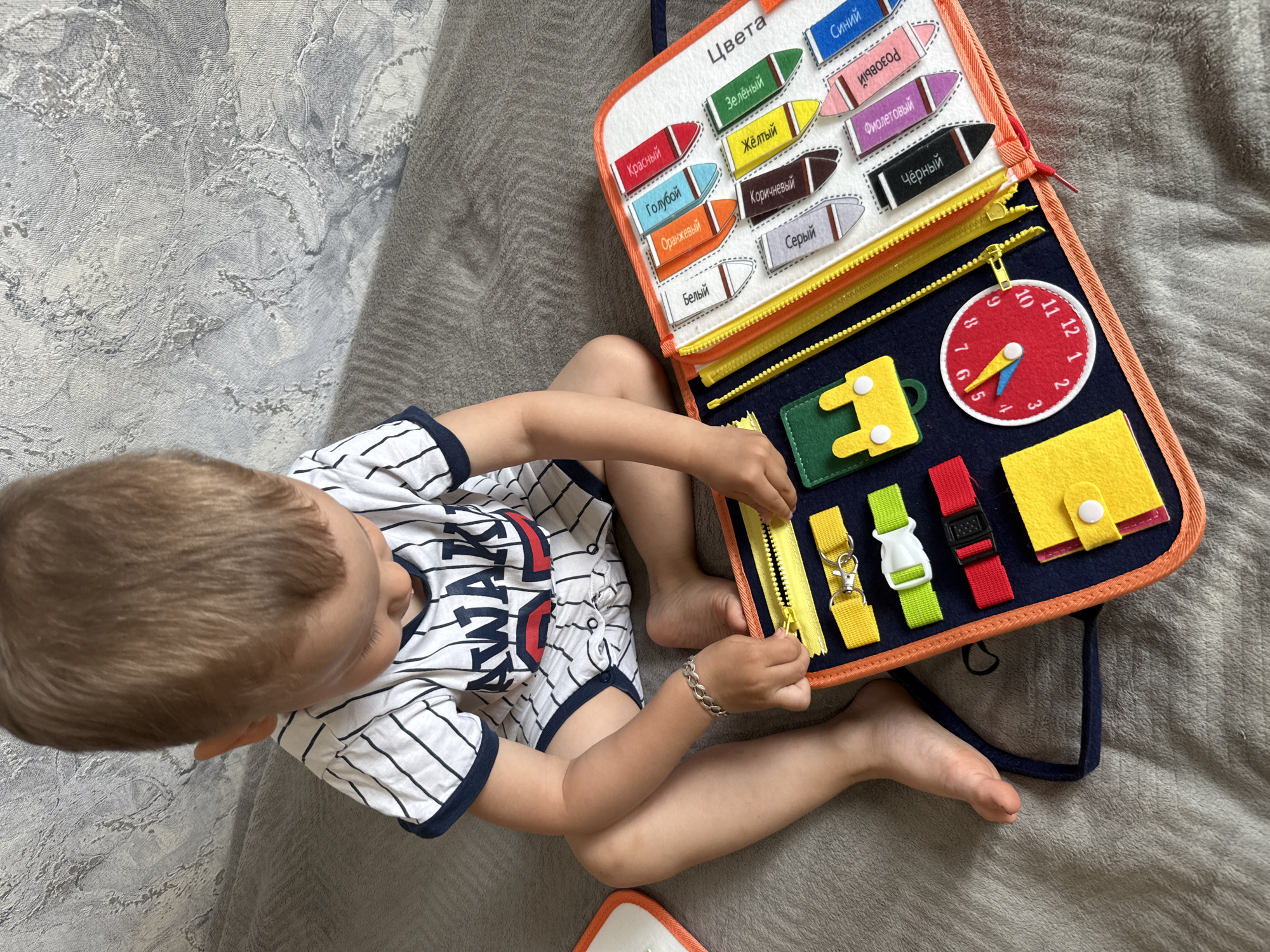 A young child is playing with a busy board toy on a gray surface that appears to be a bed or blanket. The child is wearing a white and navy striped t-shirt with a baseball-themed design and has a bracelet on their wrist. The busy board is open and features various tactile activities such as zippers, buckles, buttons, and a clock face. One side of the board has color names written in Russian next to corresponding colored pencils. The child is actively manipulating a yellow zipper on the busy board. Based on visual cues, the image appears to be taken indoors in a domestic setting. There is no information in the image that suggests the location is in the city of Rusanovo, Russia.