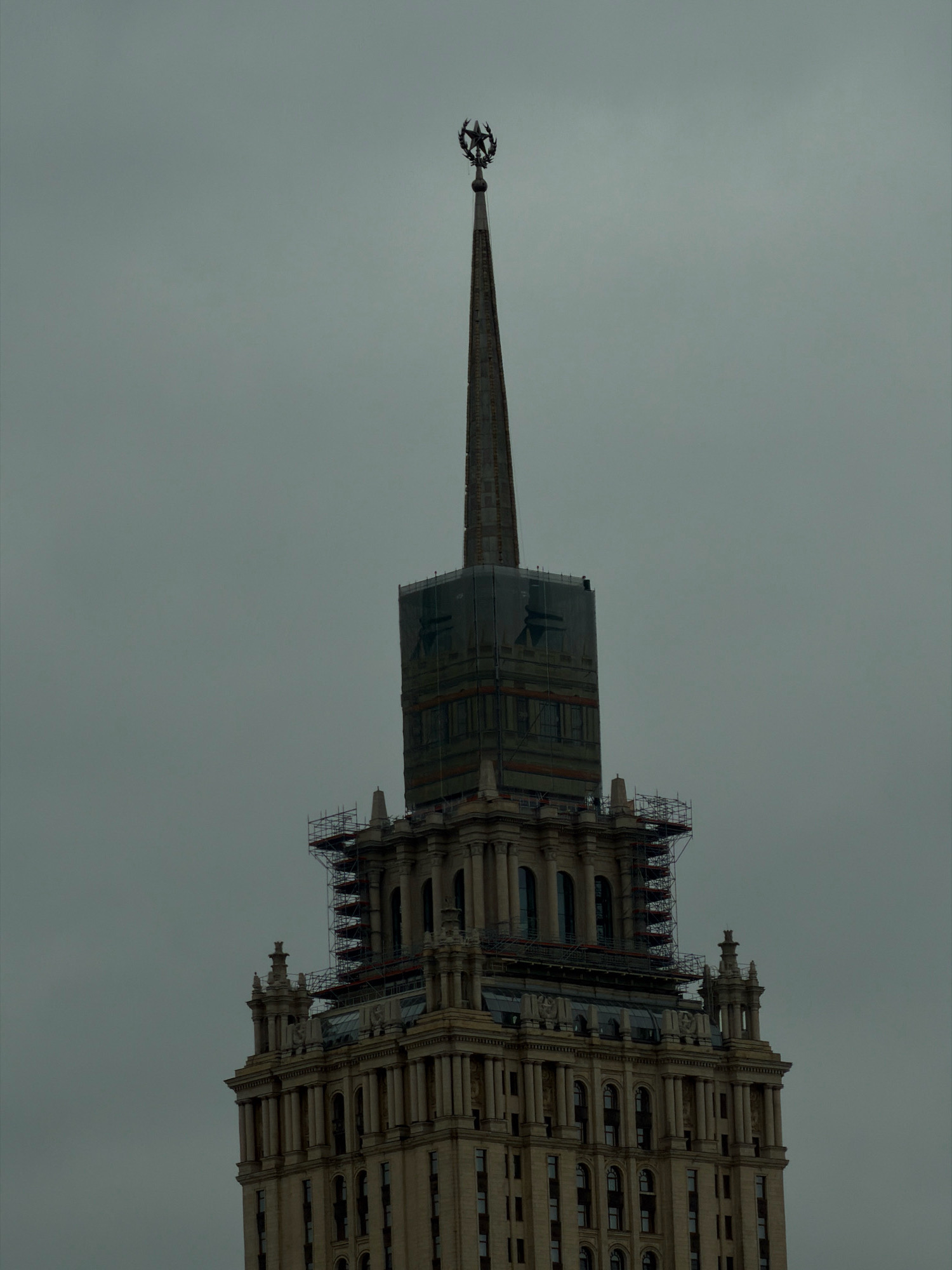 The image shows the spire of a tall, ornate building, likely in Moscow, Russia, due to its distinct architectural style and the star emblem at the very top. The building appears to be made of light-colored stone and features classical architectural elements such as columns and decorative carvings. Scaffolding is visible on the upper sections of the building, suggesting ongoing renovation or maintenance work. The sky is overcast and gray, contributing to a somber atmosphere. The overall impression is one of grand architecture under a cloudy sky, with signs of human intervention for preservation.