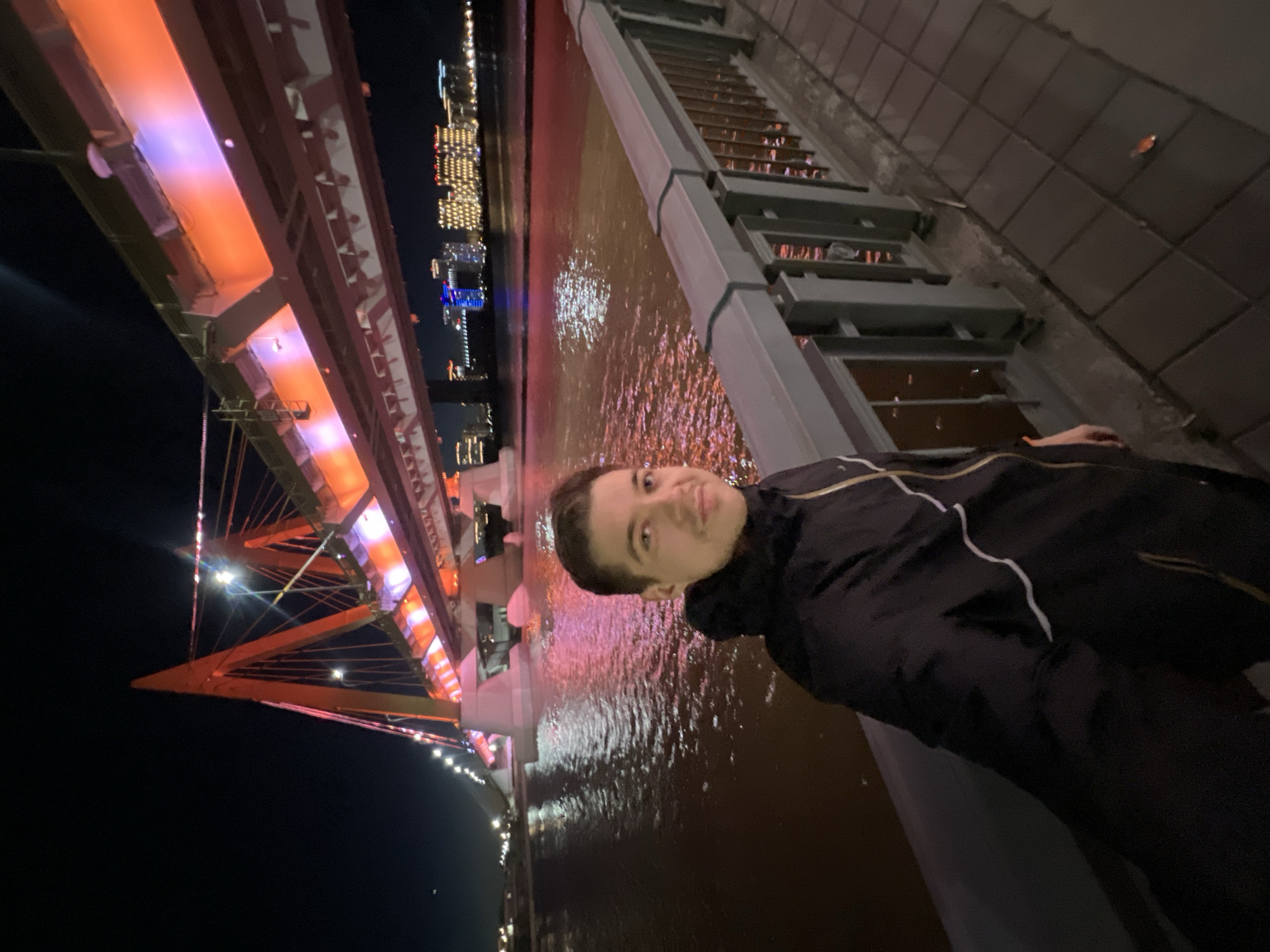 A male individual is shown in a vertically-oriented photograph, likely a selfie. The person is fair-skinned with short brown hair, wearing a dark jacket with light-colored trim. The background features the cityscape of Kazan, Russia, at night. The Millennium Bridge, illuminated with orange and purple lighting, is a prominent structure in the backdrop. The water below the bridge reflects the city lights. A bridge walkway with a grey metal railing is visible near the subject.