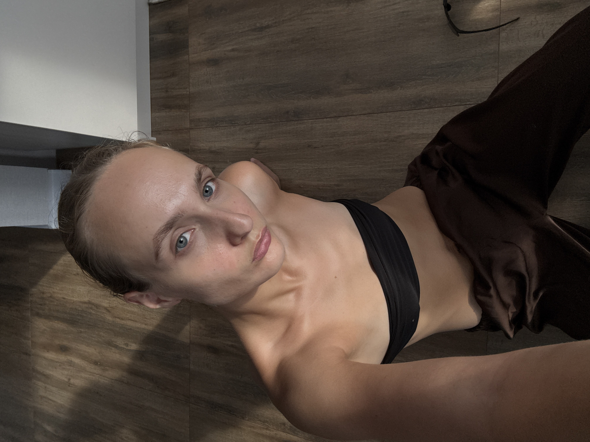 A high-angle, medium shot captures a young woman with light skin and blonde hair pulled back, sitting on a wooden floor indoors. She is wearing a black bandeau top and brown satin pants. Her head is tilted, and she is looking directly at the camera with a neutral expression. The lighting casts shadows on the floor, creating a dramatic effect. The background consists of the wooden floor and a sliver of a white wall. The scene appears to be in a residential setting in the city of Podshivalovo, Russia.
