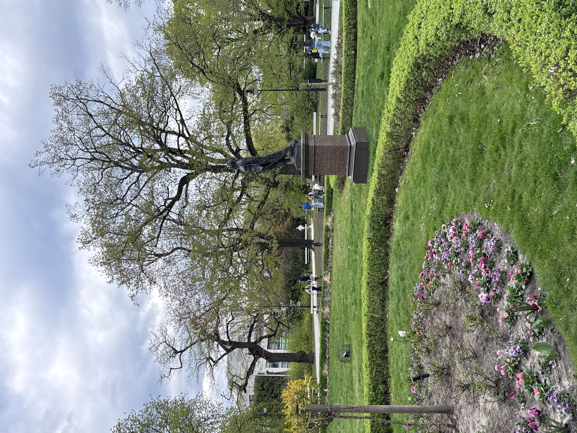 The image shows a park scene in Warsaw, Poland. In the center of the image is a statue of a figure on a pedestal. Large trees with early spring foliage provide a backdrop to the statue. Several people are visible on a path behind the statue, some sitting on benches. The foreground features a well-manicured lawn with neatly trimmed hedges. A flower bed with pink and purple flowers is located on the right side of the frame. A wooden fence post and a tree support are visible at the bottom. The sky is overcast with a mix of light blue and gray clouds. Buildings can be seen in the distance on the left.