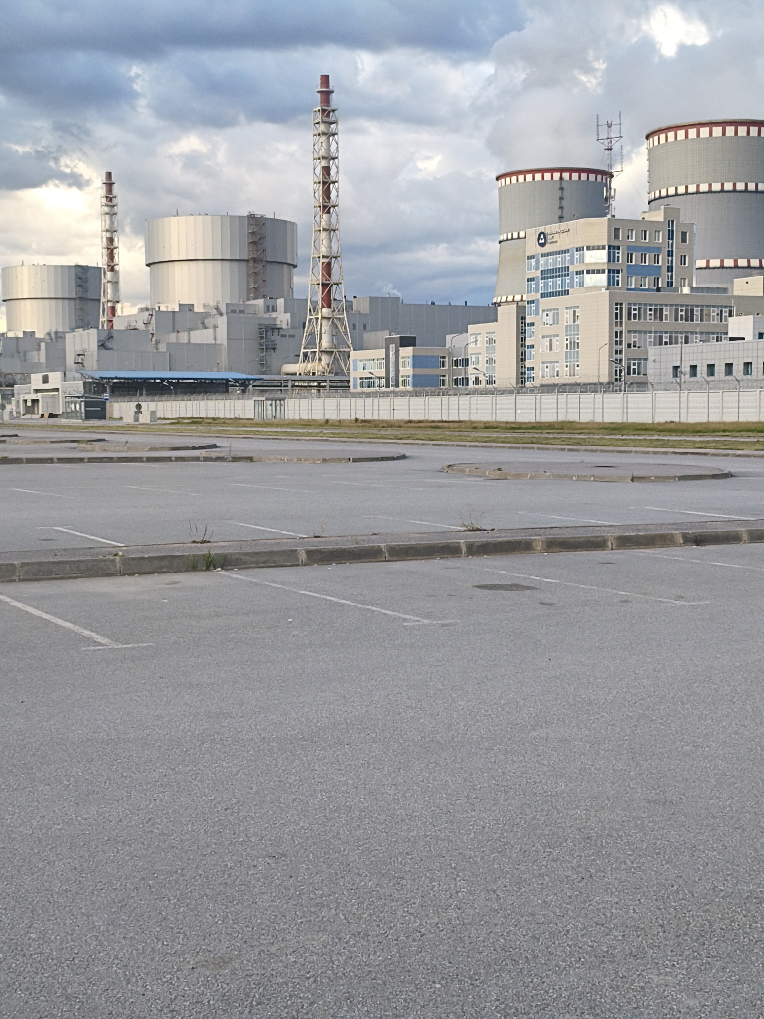 The image shows a view of the Leningrad Nuclear Power Plant in Sosnoviy Bor, Russia. In the foreground, there is an empty asphalt parking lot with white painted lines marking the parking spaces. A low concrete curb separates the parking lot from a strip of grass and then a security fence. Behind the fence, the industrial complex of the nuclear power plant is visible. The complex includes several large, cylindrical cooling towers, one of which is emitting steam or water vapor. There are also various other industrial buildings and tall, lattice-like structures with red and white banding, which are likely chimneys or vents. The sky above is partly cloudy with a mix of grey and white clouds. The overall scene is one of industrial infrastructure under a cloudy sky.