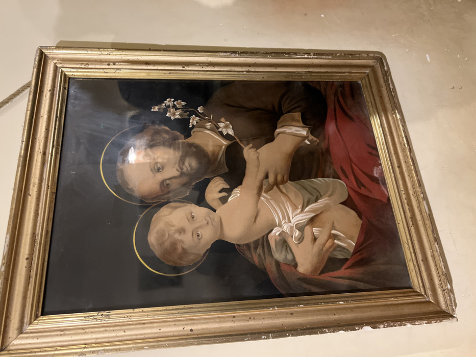 The image is a painting in a gold-colored frame depicting Saint Joseph holding the Christ Child. The painting is hanging on a white wall. The scene appears to be set indoors. The figures in the painting are depicted against a dark background. Saint Joseph is on the right side of the painting, holding the Christ Child on his left arm. The Christ Child is wearing a white cloth around his waist. Saint Joseph has a dark beard and mustache and is wearing a dark blue robe. The Christ Child is looking straight ahead with a serious expression. Saint Joseph is looking off to the left side of the painting. The painting appears to be old and slightly faded. The image was taken at the location with coordinates (latitude: 40.852, longitude: 14.257) which is in the city of Napoli, Italy.
