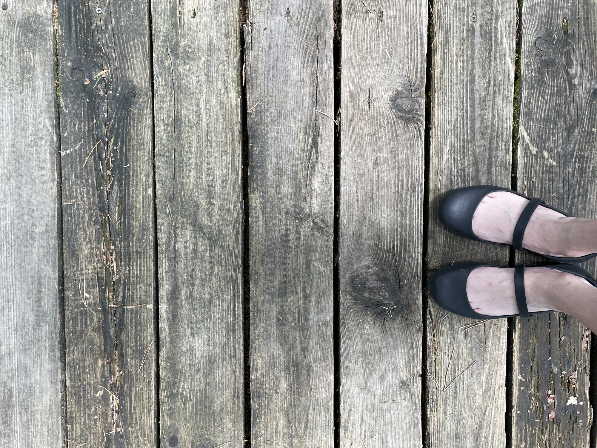 An overhead shot shows a person's feet wearing black ballet flats standing on a weathered wooden deck. The deck is made of vertical wooden planks with visible grain, knots, and cracks, indicating age and outdoor exposure. Small pieces of debris, such as pine needles and bits of dirt or moss, are scattered across the surface. The feet are positioned on the right side of the frame, with one foot slightly in front of the other. The shoes are simple, flat, and black, with a strap across the instep. The overall scene suggests a casual outdoor setting, possibly a backyard, park, or boardwalk. The lighting is even, suggesting it could be daytime, with no strong shadows or glare. No text is visible in the image.