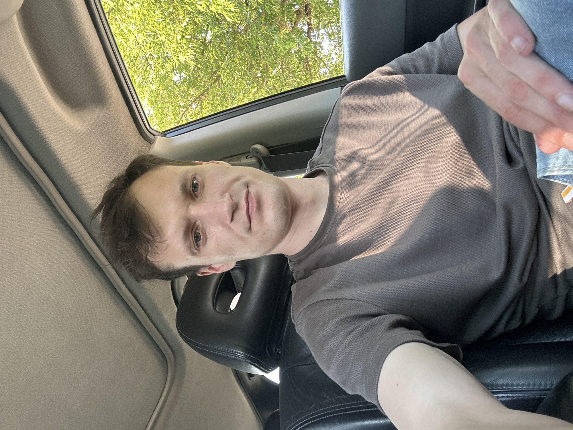 A young Caucasian man is sitting in the driver's seat of a car, looking directly at the camera. He has short, dark brown hair and fair skin. He is wearing a grey, short-sleeved t-shirt. The interior of the car is visible, including the black leather seats and a sunroof. Outside the car window, a tree with bright green leaves is visible, suggesting a sunny day. The image appears to be taken in Makiv, Ukraine, likely during daylight hours.