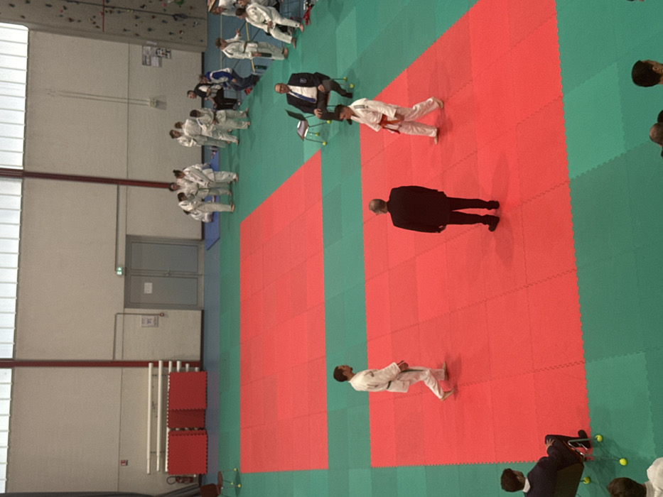 This image depicts an indoor gymnasium in Brumath, France, set up for a martial arts event, likely judo. The floor consists of interlocking green and red mats. Two large, rectangular red mat sections are centrally placed on the green matting. The gymnasium walls are light-colored, with a band of translucent panels or windows near the ceiling. A climbing wall with various holds is visible on the right side. Double grey doors are present in the far wall, and a fire extinguisher is mounted on the left wall near some horizontal bars.
In the foreground, on the lower red mat, three individuals are present: a barefoot male in a white judogi with a black belt is walking from right to left; a male in a dark suit stands with his back to the viewer, facing away; and a male in a white judogi with an orange belt, wearing white footwear, walks from left to right.
To the right of the foreground red mat, a male official in a dark jacket, white shirt, and blue tie with an insignia is seated on a chair, observing the mat. On the far right, adjacent to the climbing wall, a group of approximately ten to twelve individuals in white judogis (some with various belt colors) are standing and sitting on chairs. In the lower-left foreground, a male in a dark jacket is seated on a chair, observing the scene, and other individuals are partially visible at the bottom edge. Empty chairs with yellow leg tips are positioned around the periphery.