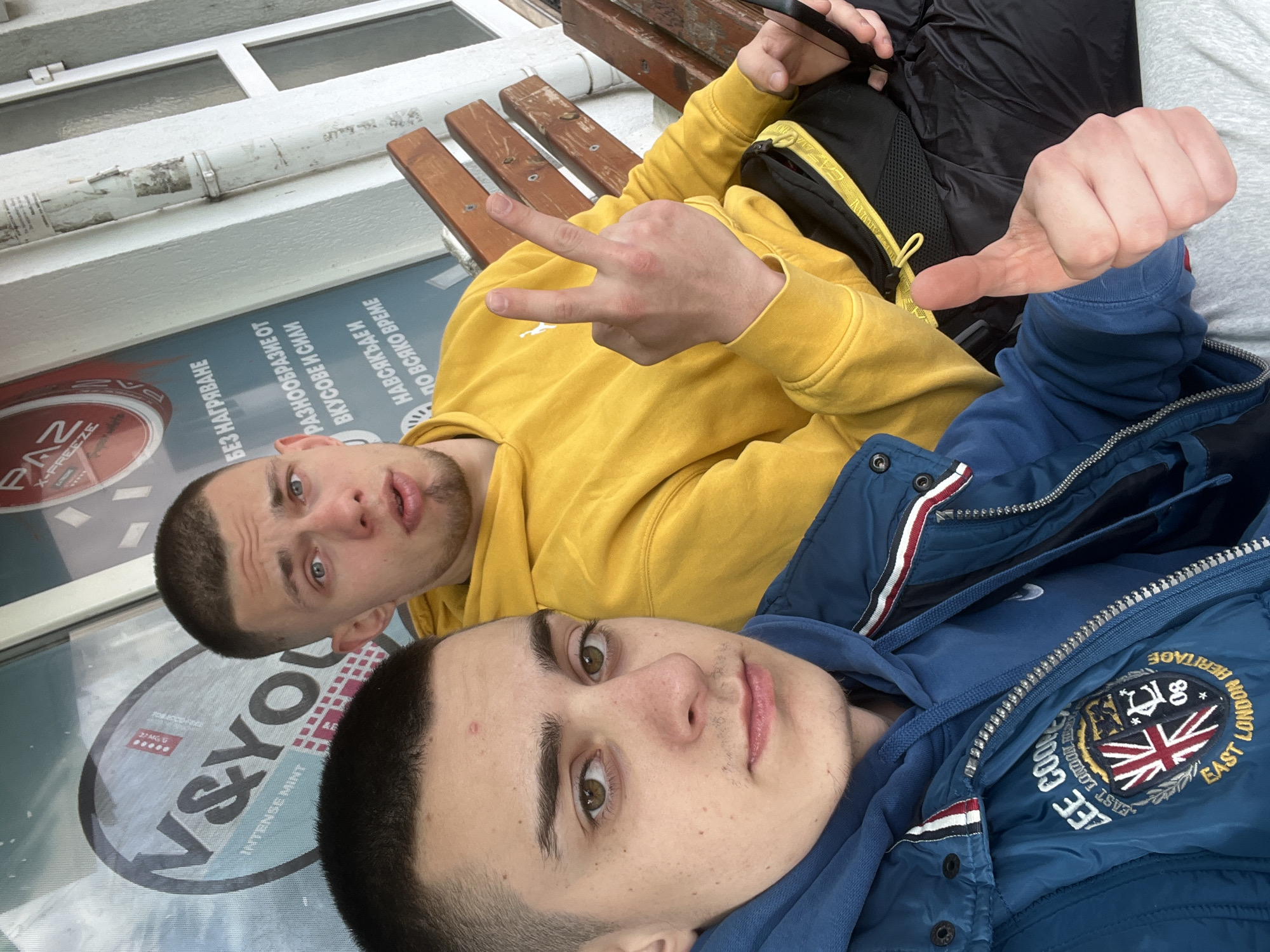 Two young men are lying on a bench in Burgas, Bulgaria, in front of a storefront window, taking a selfie. The man in the foreground is wearing a blue hoodie and a blue jacket with a "LEE COOPER EAST LONDON HERITAGE" patch featuring a Union Jack. The man behind him is wearing a yellow hoodie and making a peace sign with one hand and pointing with the other. The storefront window has a "V&YOU INTENSE MINT" logo and other text in Bulgarian, including the words "PAZ X-FREEZE" and "БЕЗ НАГРЯВАНЕ". The bench is made of wood and metal.