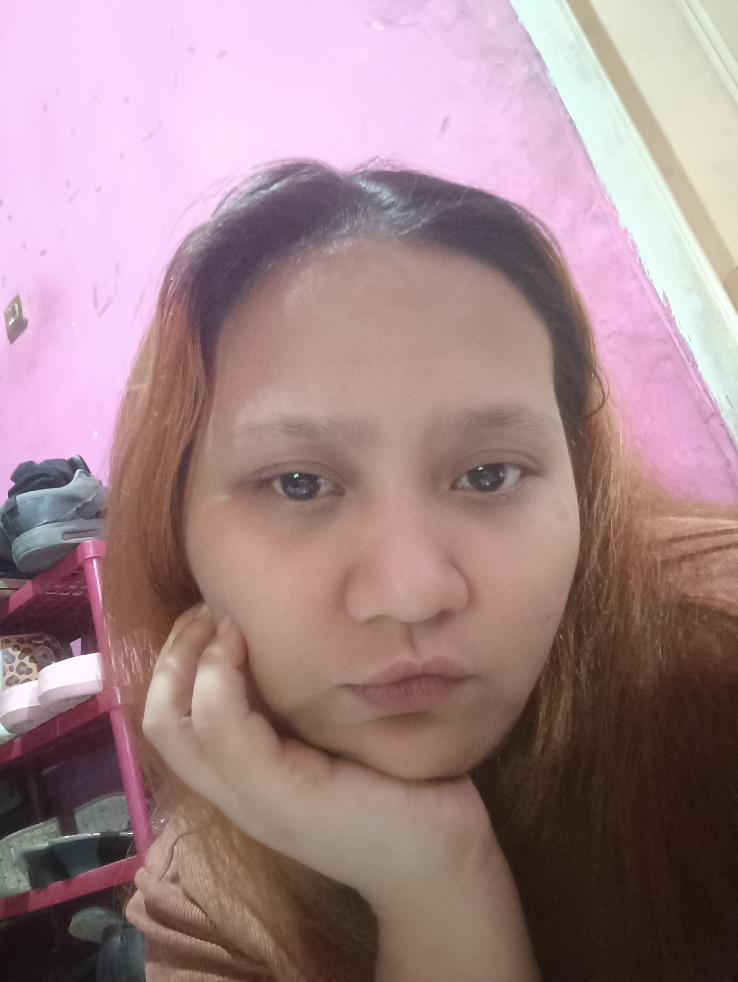 The image is a close-up, selfie-style portrait of a young woman with auburn hair, resting her chin on her hand. She has light brown eyes and is looking directly at the camera. The background is a pink wall with some scattered dark marks and a white trim on the right side. To the left of the woman, a pink plastic shelving unit is visible, holding a pair of grey sneakers and other items. The lighting appears to be indoor, with a slight cast. The overall scene suggests a personal, casual setting, possibly within a residential space in Depok, Indonesia, though no specific landmarks or urban indicators are present in the frame.