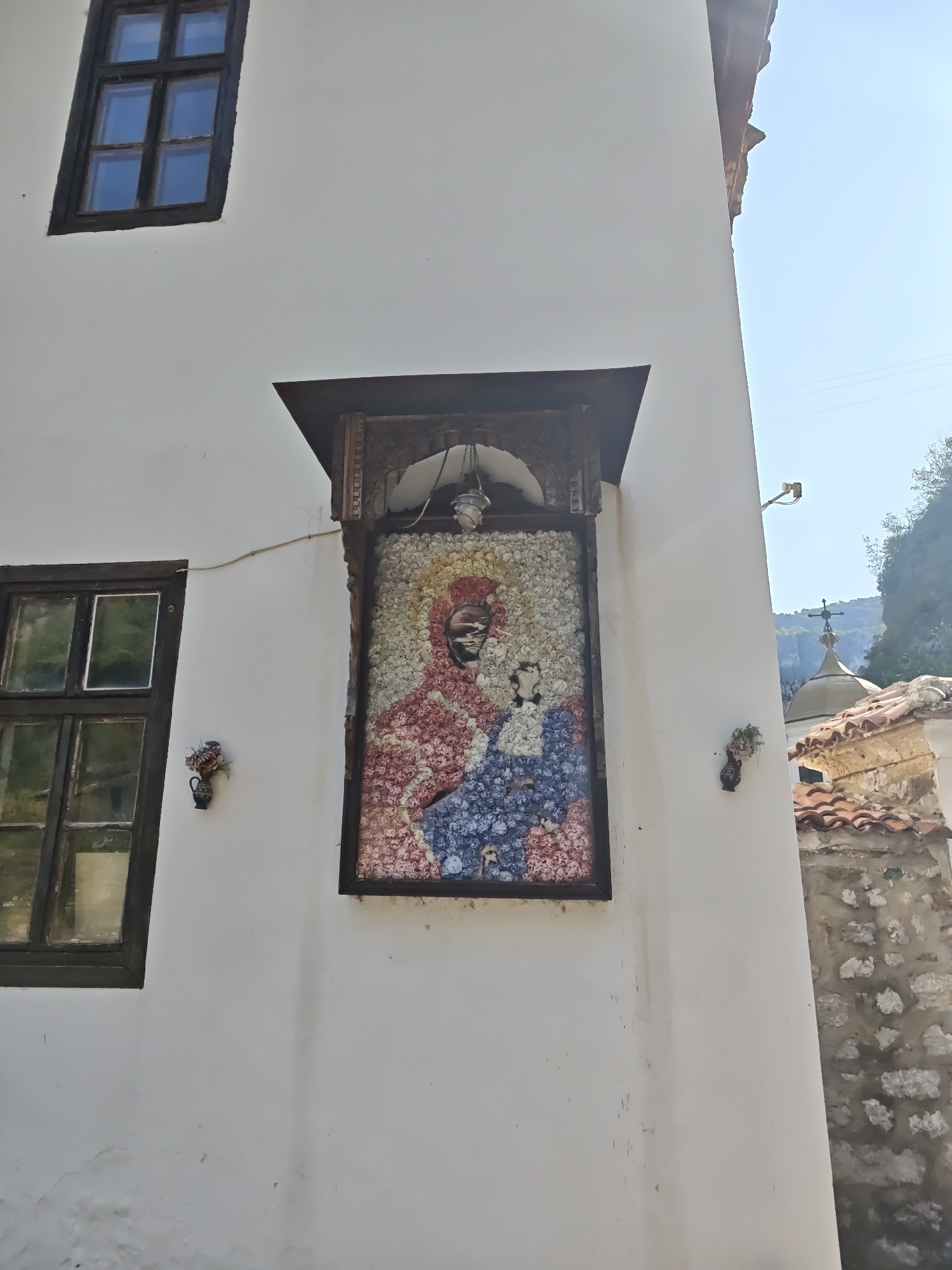 The image depicts a section of the exterior wall of a building in the city of Lyutibrod, Bulgaria. The building has white plastered walls and dark wooden window frames. A prominent feature on the wall is a mosaic artwork, framed in dark wood and set within a decorative alcove. The mosaic depicts a religious figure, likely the Virgin Mary holding the Christ child, rendered in colorful tiles of red, blue, and white. The figures' faces are detailed with smaller, darker tiles. Above the mosaic, a carved wooden canopy is visible. To the left of the mosaic, a dark-framed window with multiple panes is present. Small, dark vases with artificial flowers are attached to the wall on either side of the mosaic. In the background, a portion of a red-tiled roof and a stone wall are visible. Further in the distance, the dome and cross of a church are silhouetted against a clear blue sky, with lush green trees and rocky cliffs framing the scene. The overall impression is one of a historic religious site, possibly a monastery or church, bathed in sunlight.