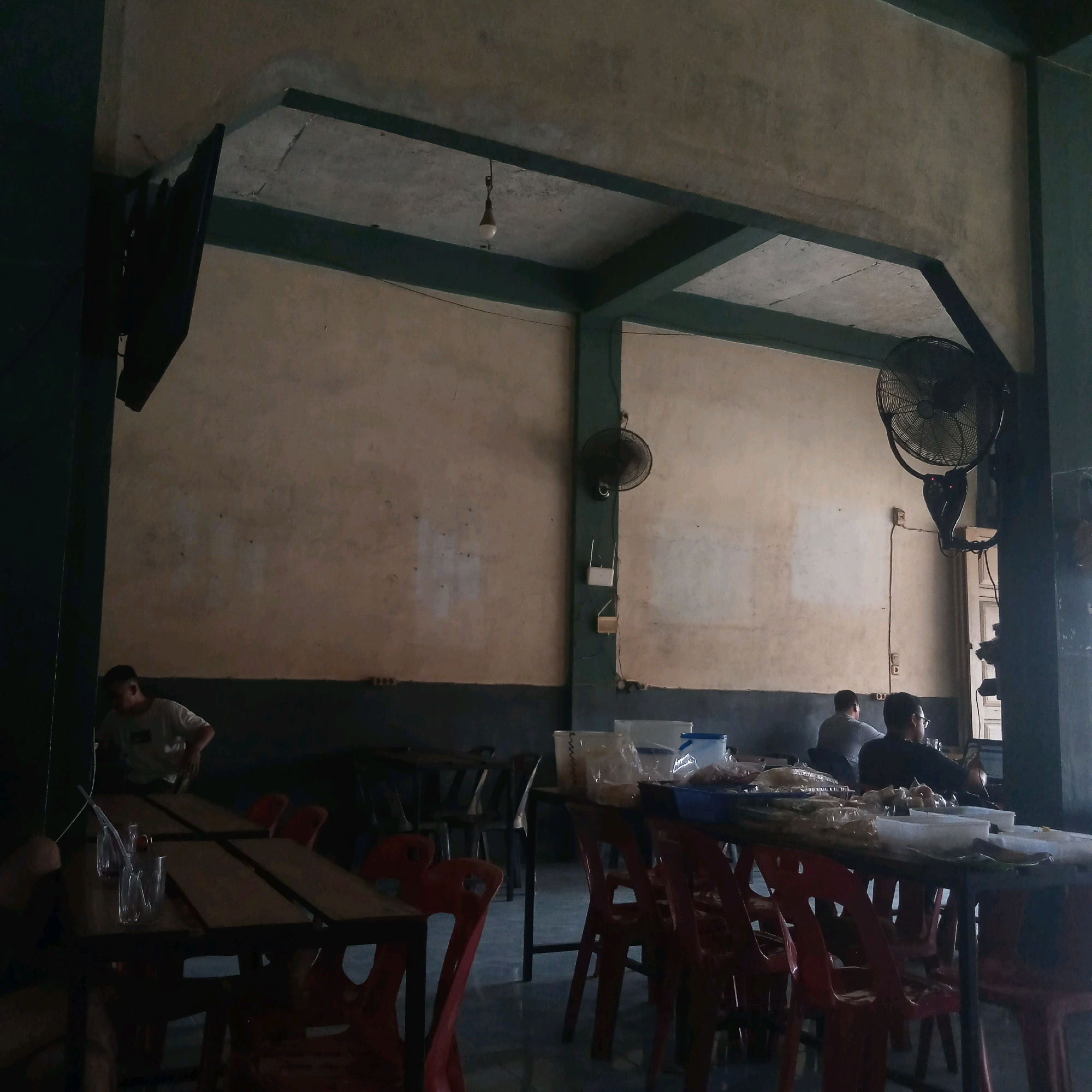 The image depicts the interior of a dimly lit eatery, likely a warung or simple restaurant, in Grong-Grong, Indonesia. Several wooden tables with dark metal frames are arranged in the dining area, many of them occupied by red plastic chairs. On the tables, there are various items including plastic cups, straws, plastic-wrapped food items, and containers of food.
A young man wearing a light-colored t-shirt is seen standing near a table on the left side of the frame, appearing to be engaged with items on the table. Further back, several other individuals are seated at tables, their faces mostly obscured or turned away from the camera. The walls of the establishment are a light, mottled color, with a darker band running along the lower portion. Overhead, exposed beams painted in a dark green color support the ceiling, from which a single light bulb hangs. Two industrial-looking oscillating fans are mounted on the walls, one on the left and another on the right side of the room, indicating the need for ventilation and cooling in the local climate. A flat-screen television is mounted on the wall on the far left, and a wifi router is visible mounted on a dark green pillar on the right. The flooring appears to be tiled. The overall impression is a functional, casual dining space.