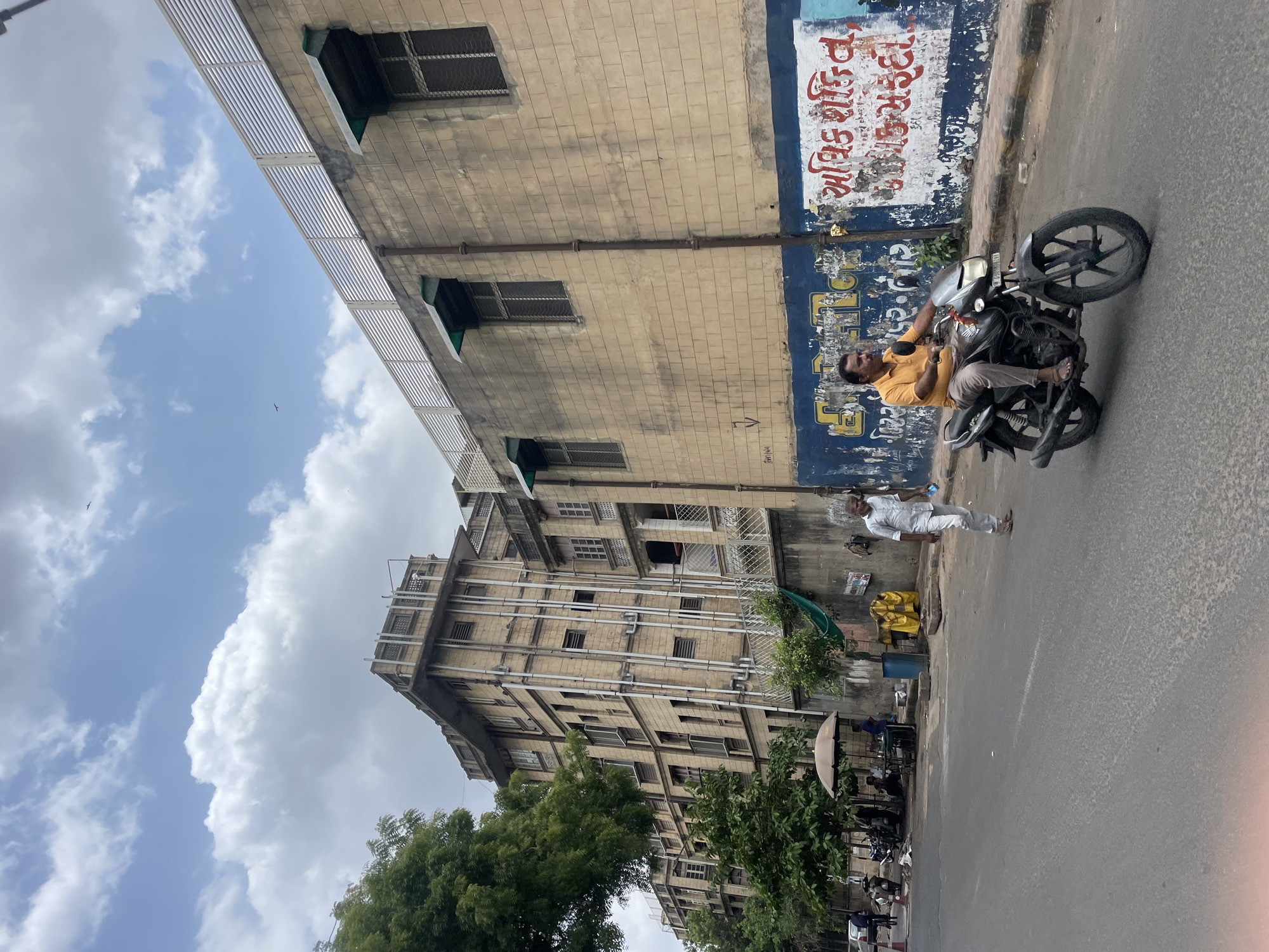 The image captures a street scene in Ahmedabad, India, with a focus on a man riding a motorcycle on a paved road. In the background, a multi-story building with a beige brick facade stands prominently. The building features several windows, some with green awnings, and external pipes running vertically along its side. A white slatted fence is visible on the rooftop. To the left of this building, another older, taller building with a similar brick construction and numerous windows is partially visible, with a large tree in the foreground. The road has some subtle textural variations. On the wall of the beige building, there is graffiti in a blue and white color scheme. Another man in a light-colored shirt and pants is walking on the sidewalk to the right of the motorcycle. Further down the street, a small roadside stall with an umbrella can be seen, with several motorcycles parked nearby. The sky above is partly cloudy with a blue hue.
