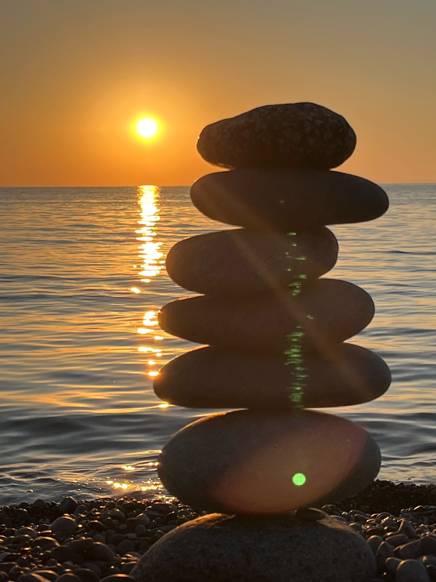 A vertical stack of six smooth, grey stones is positioned on a pebble beach at the edge of a calm body of water. The sun, a bright orange orb, is low on the horizon, casting a golden glow across the sky and reflecting a shimmering path on the water's surface. The background shows a vast expanse of orange-tinted sky meeting the horizon of the water. There are no people or animals present in the image. The scene appears to be a natural, tranquil setting, likely during sunset. The location is Kobuleti, Georgia.