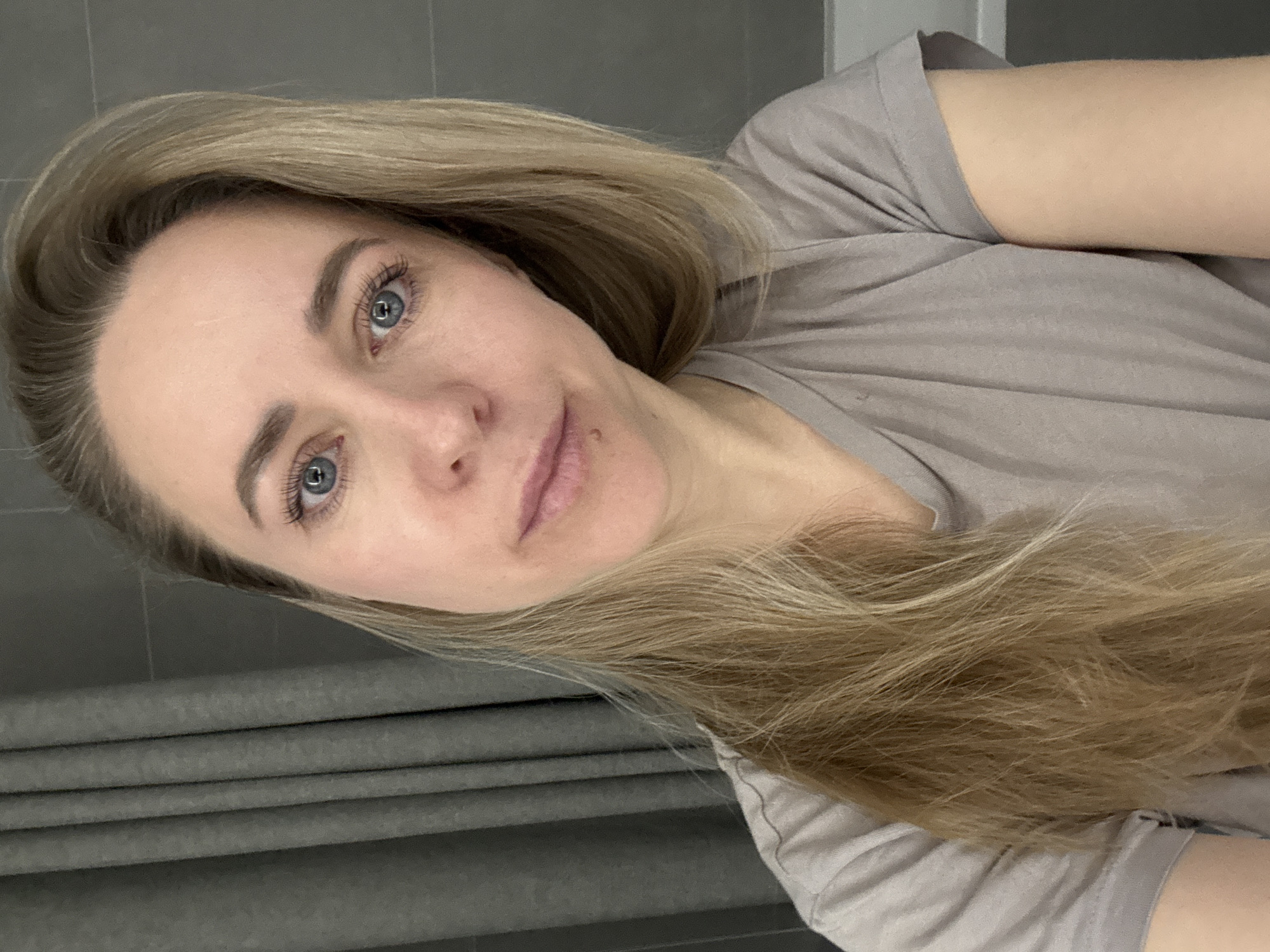 A woman with blonde hair and blue eyes is facing the camera. She is wearing a gray t-shirt. The background is a gray wall. There is no evidence indicating that this image was taken in Chaikovsky, Russia.