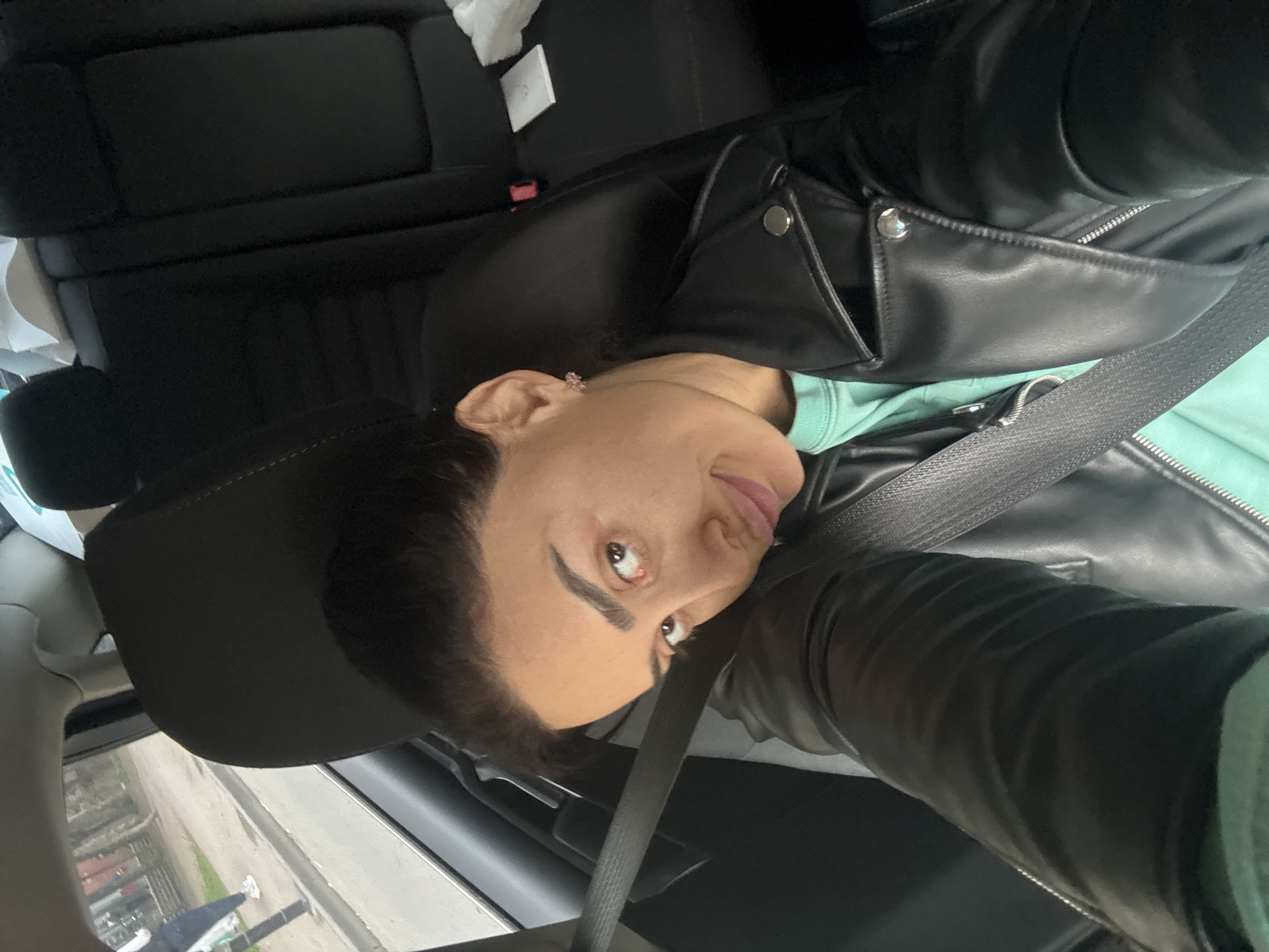 A selfie captures a person sitting in the passenger seat of a car, with the image rotated ninety degrees clockwise. The subject is a woman with dark hair pulled back, expressive brown eyes, and well-defined eyebrows, looking towards the camera with a subtle, slight smile. She wears a black faux leather jacket over a mint green top, and a seatbelt crosses her chest. A small, light pink stud earring is visible in her right ear.
The car's interior features black seats and upholstery. In the background, on the rear seat, a crumpled white tissue or paper item and a small white rectangular box are visible. Through the car window, a blurry view of an overcast outdoor scene can be seen, depicting what appears to be a street or pathway with some greenery and distant structures. The overall lighting suggests daytime under cloudy conditions. The scene is set in Tbilisi, Georgia.