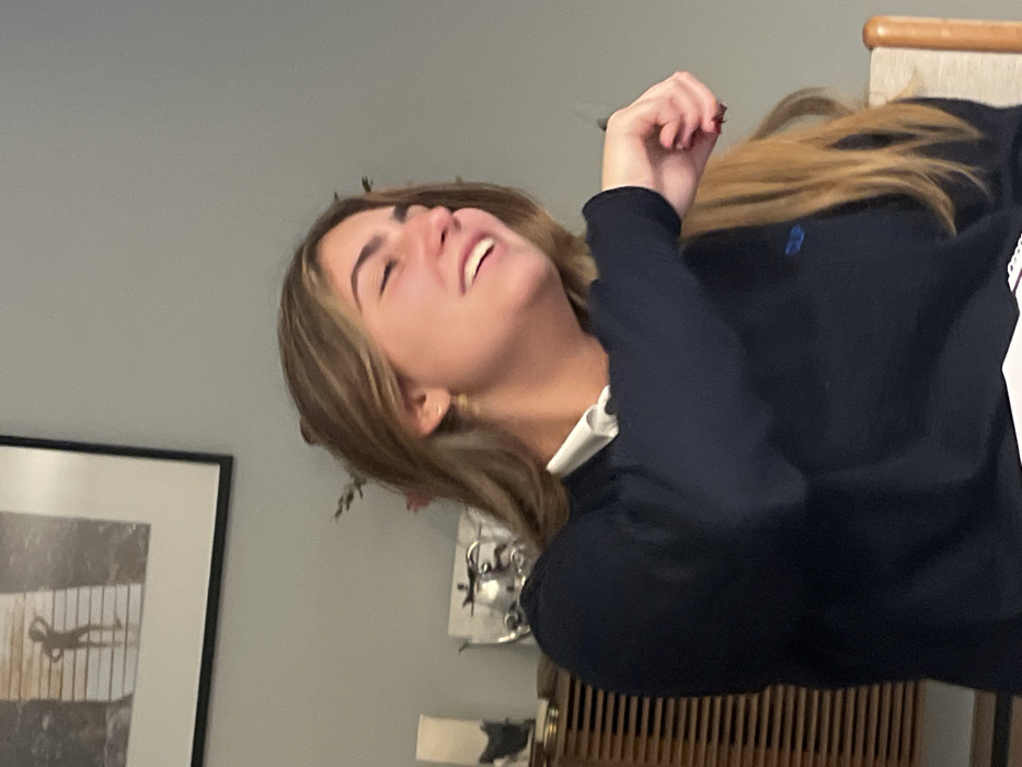 An indoor, close-up photograph features a young adult female within a domestic setting in Lordelo do Ouro e Massarelos, Portugal. The subject has light skin and long, light brown hair, captured mid-laugh or strong smile with her eyes nearly closed and mouth open. She is wearing a dark blue long-sleeved sweater over a white collared shirt; a small blue embroidered logo is visible on the sweater's left chest. Small gold-colored hoop earrings are present, and her fingernails display dark red polish. Her right arm is bent and raised, with the hand partially clenched. The background reveals a plain light gray wall. In the upper left, a black-framed picture depicts a silhouetted figure on a pier. Below it, a wooden cabinet with horizontal slat detailing holds decorative objects, including a silver-toned vessel. A portion of a light-colored wooden chair back is visible in the lower right.