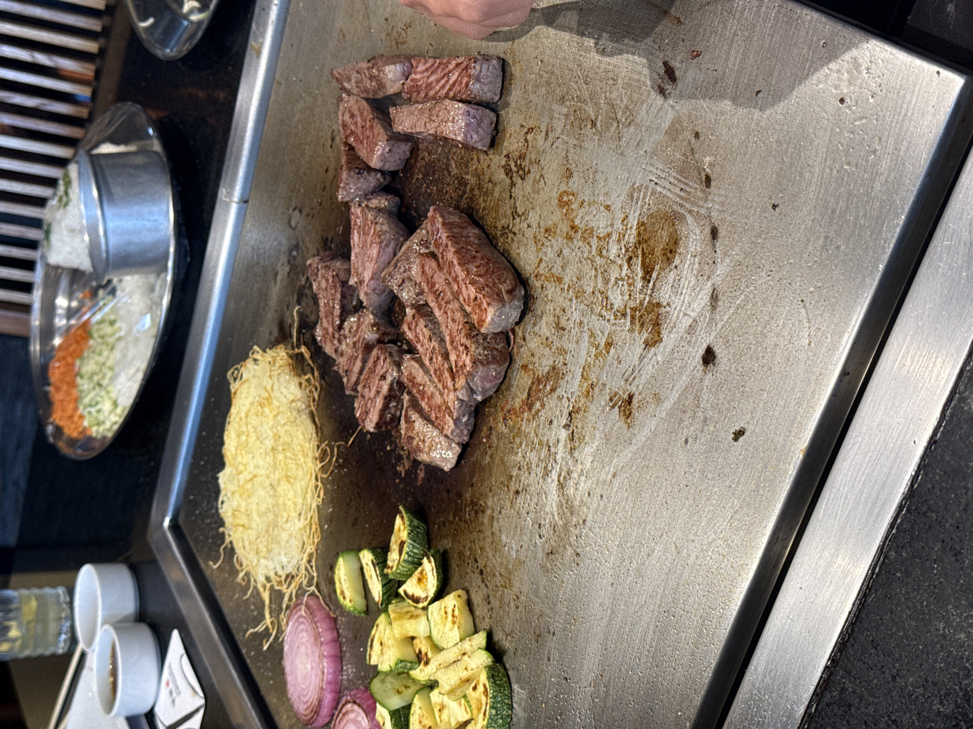 The image depicts a teppanyaki grill with various ingredients. There are pieces of cooked steak, a pile of noodles, sliced zucchini and red onion. Adjacent to the grill, there is a tray containing rice and vegetables, along with small cups and a container of dipping sauce. The location is likely a teppanyaki restaurant, possibly in Puebla, Mexico, but this is impossible to confirm from the image alone.