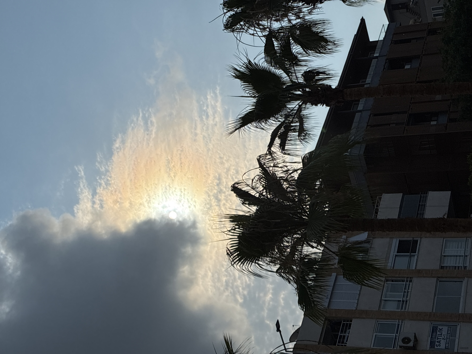 The image shows a view of a building and palm trees in Alanya, Turkey. The sun is partially obscured by clouds, creating a colorful halo effect. The building is a multi-story structure with visible windows and balconies. Several palm trees with fronds are in the foreground, silhouetted against the sky. The sky is partly cloudy with a clear blue section visible. The scene appears to be captured during daytime.