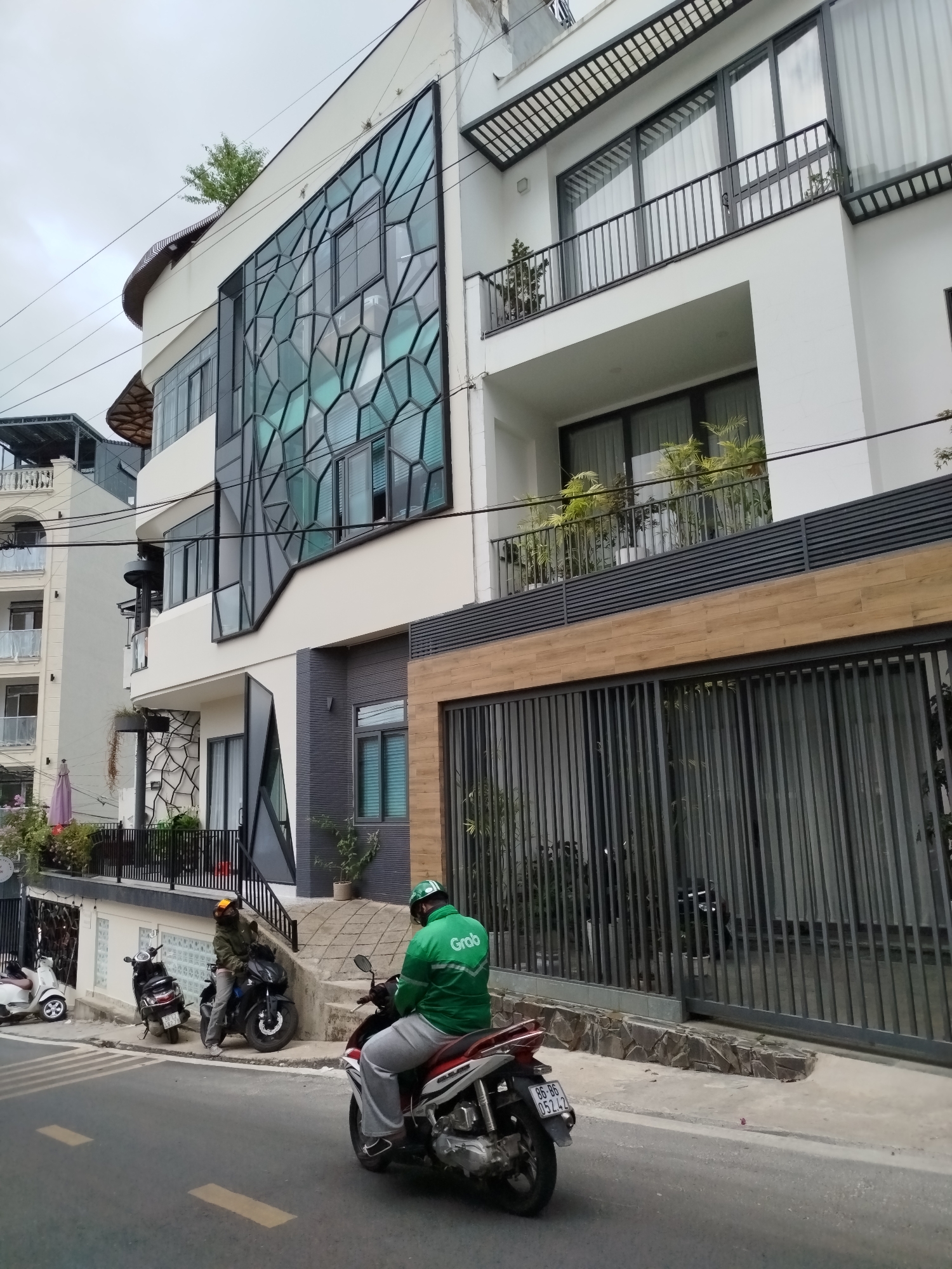 The image shows a street scene in Da Lat, Vietnam. A modern, multi-story residential building dominates the background. The building features a unique architectural design with a large geometric window pattern. A "Grab" delivery driver is on a motorcycle in the foreground, facing away from the camera. Several other motorbikes are parked on the sidewalk beside the building. The road is asphalt, with yellow lane markings. The sky is overcast. A gated fence runs along the front of the building. Adjacent to the building is another residential structure, more traditional in style.